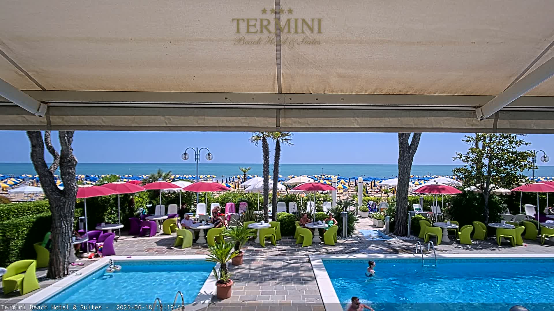 A view from under a patio awning shows a sunny outdoor pool area with colorful umbrellas and lounge chairs, overlooking a beach and calm ocean.