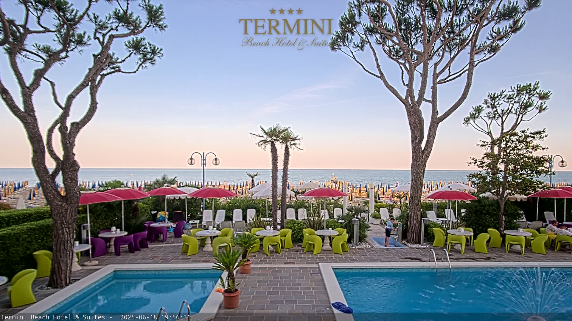 A patio with two pools overlooks a beach with numerous umbrellas under a twilight sky.