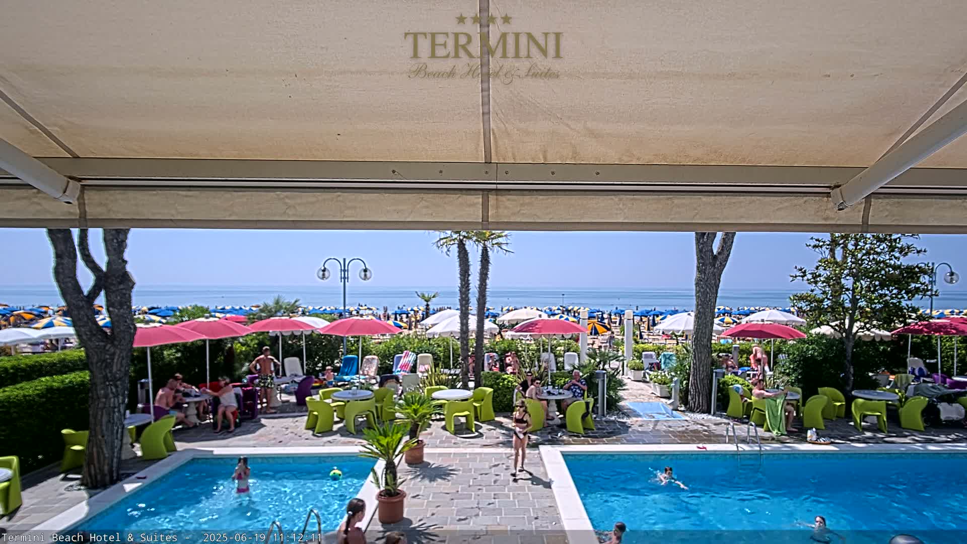 A sunny outdoor scene shows a patio with two swimming pools and people lounging around tables under umbrellas, overlooking a beach crowded with colorful umbrellas and sunbathers.