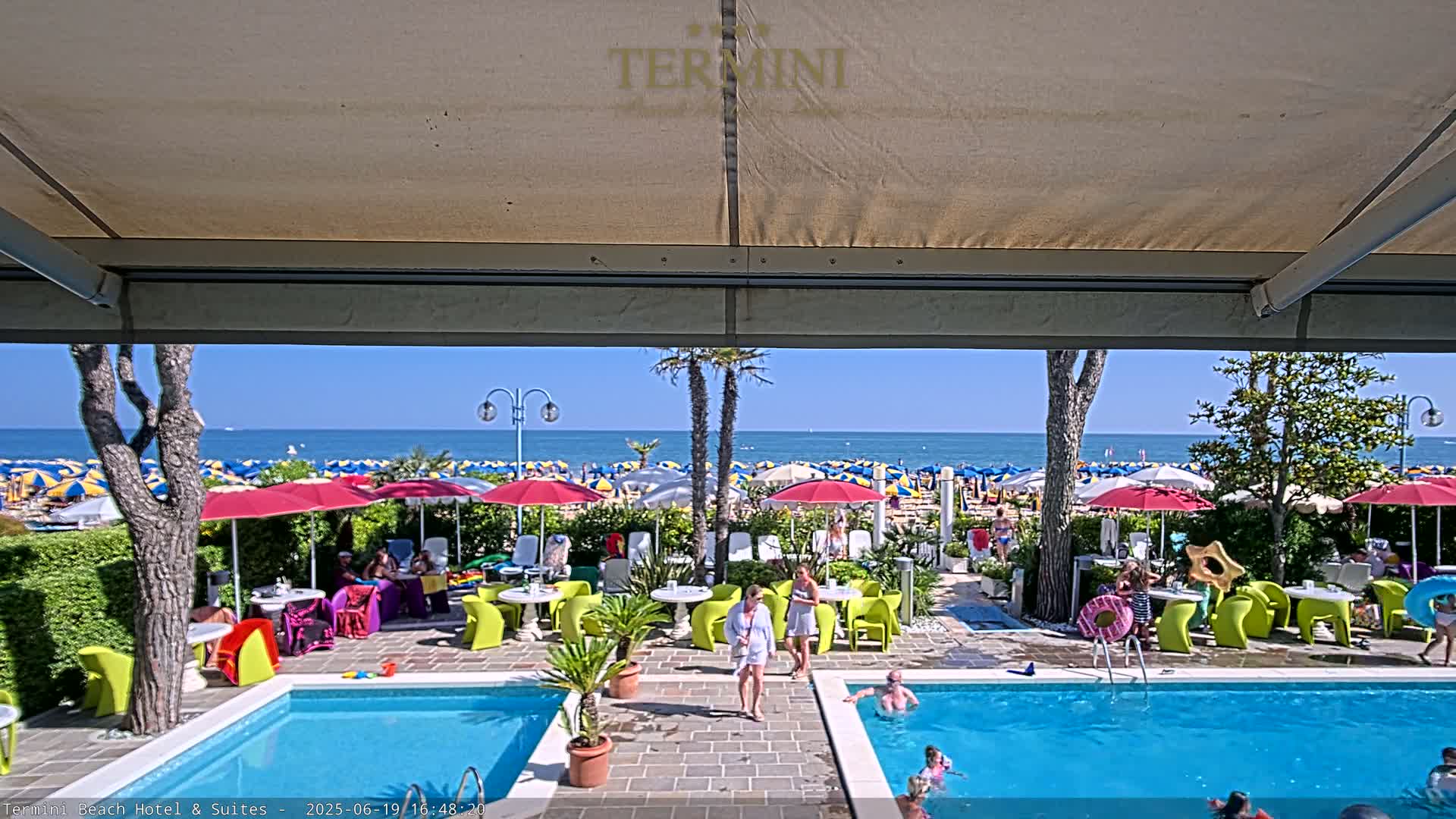 A sunny day at a beachside hotel features a pool area with people relaxing and swimming, surrounded by colorful umbrellas and lounge chairs, overlooking a beach full of more umbrellas and people.