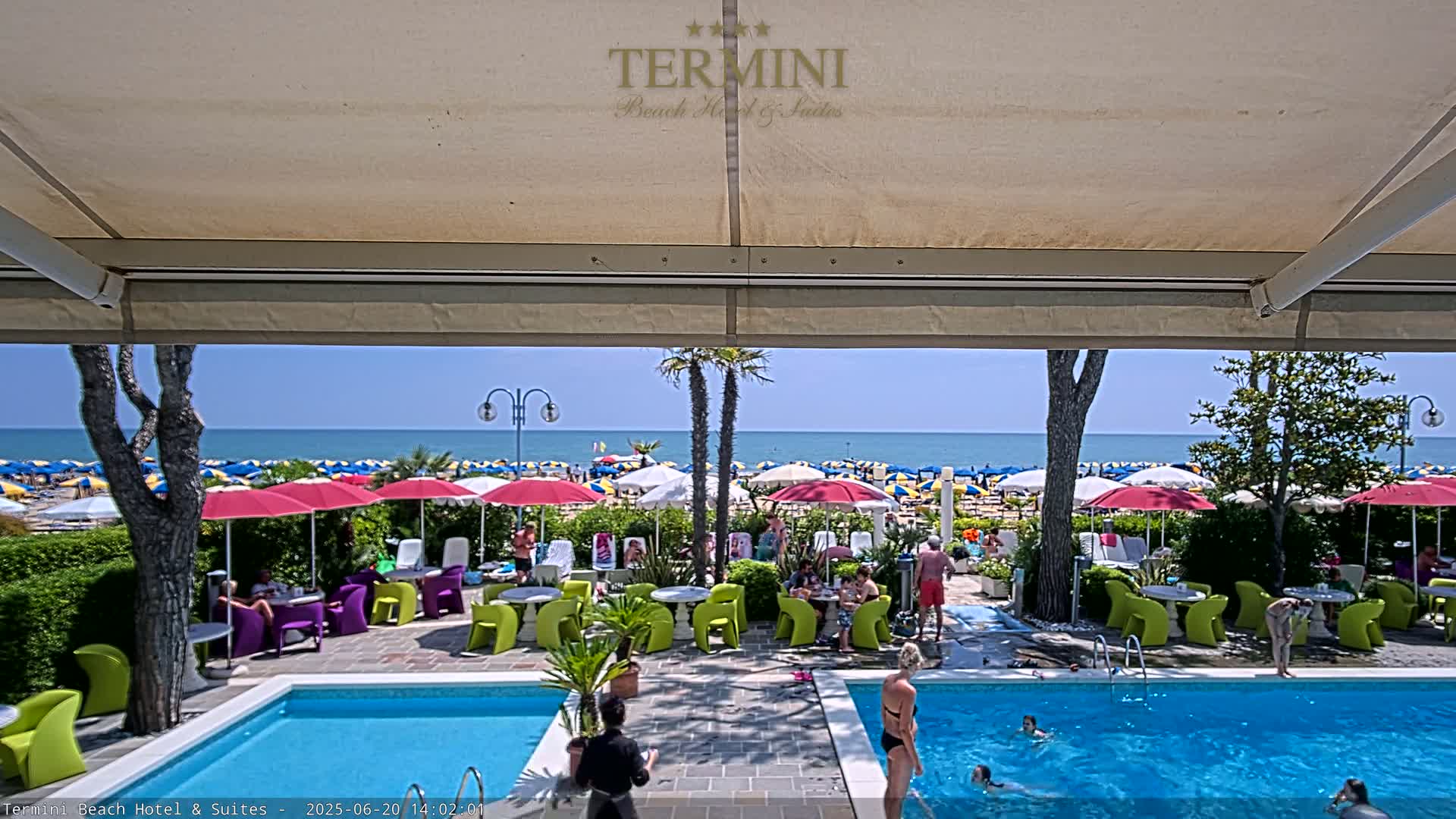 A swimming pool and patio area with brightly colored chairs and umbrellas overlook a sunny beach and calm ocean.
