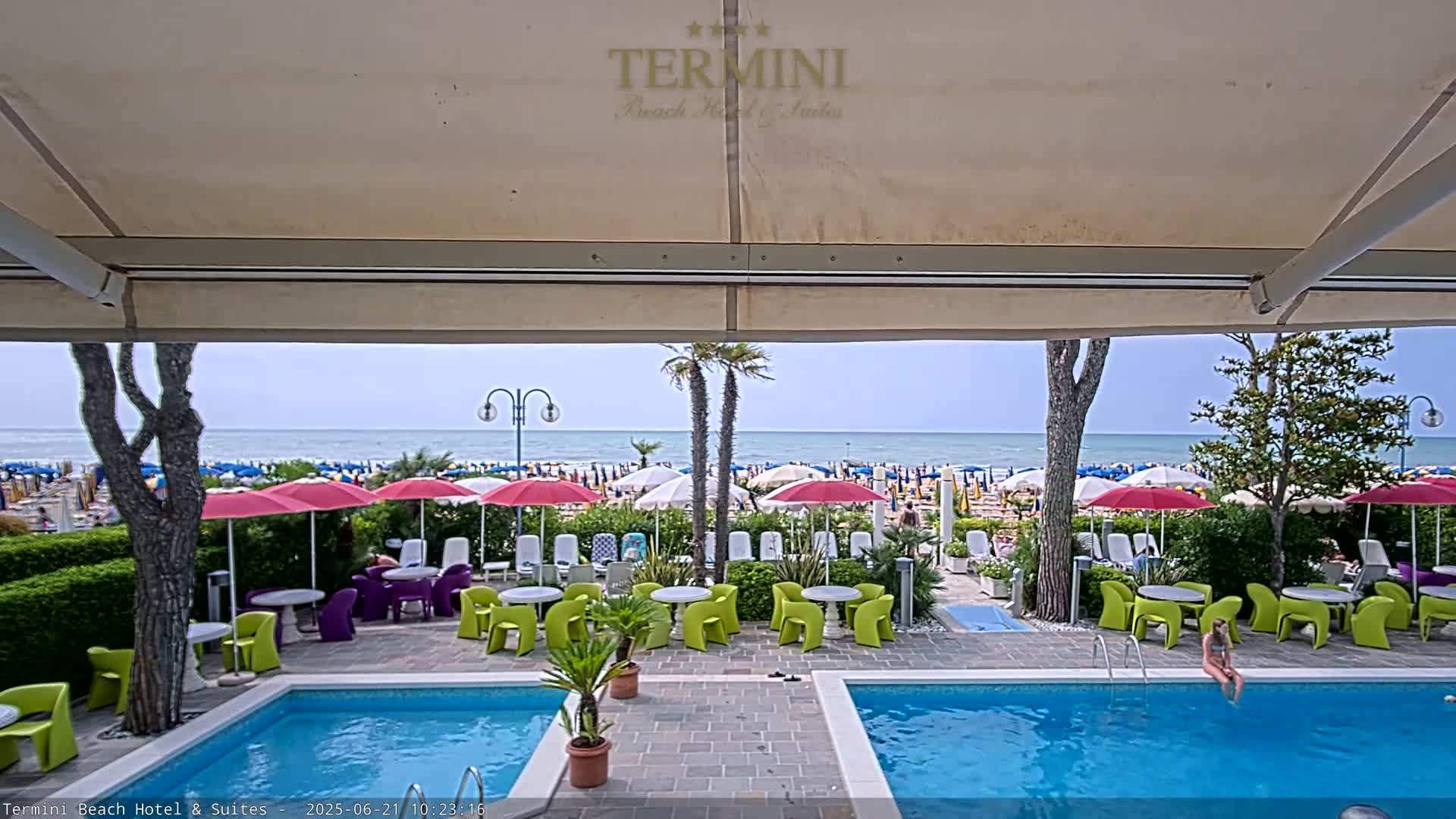 A view from under a patio awning shows a swimming pool, patio furniture, and a beach with many umbrellas and people under a mostly clear sky.