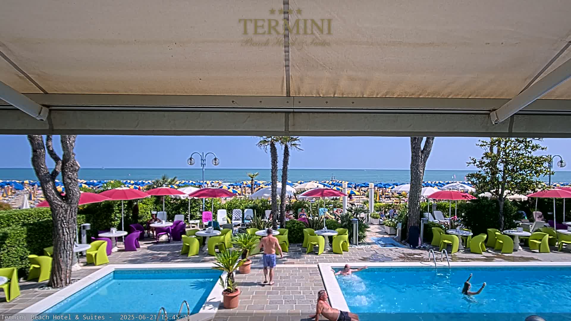 A sunny day at a resort shows a swimming pool with people swimming and sunbathing, and a beach with numerous colorful umbrellas beyond.