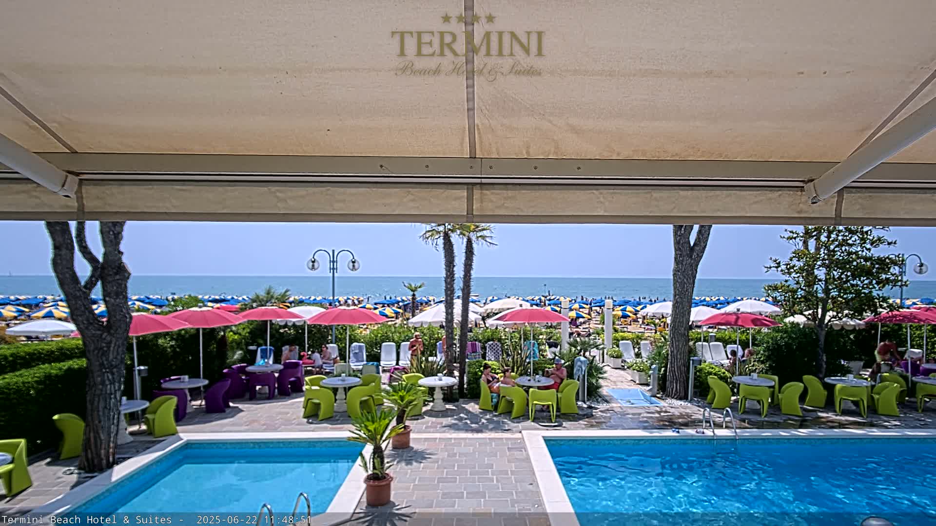 A sunny outdoor scene shows a swimming pool and patio furniture at a beachfront hotel, with a beach full of colorful umbrellas visible beyond.