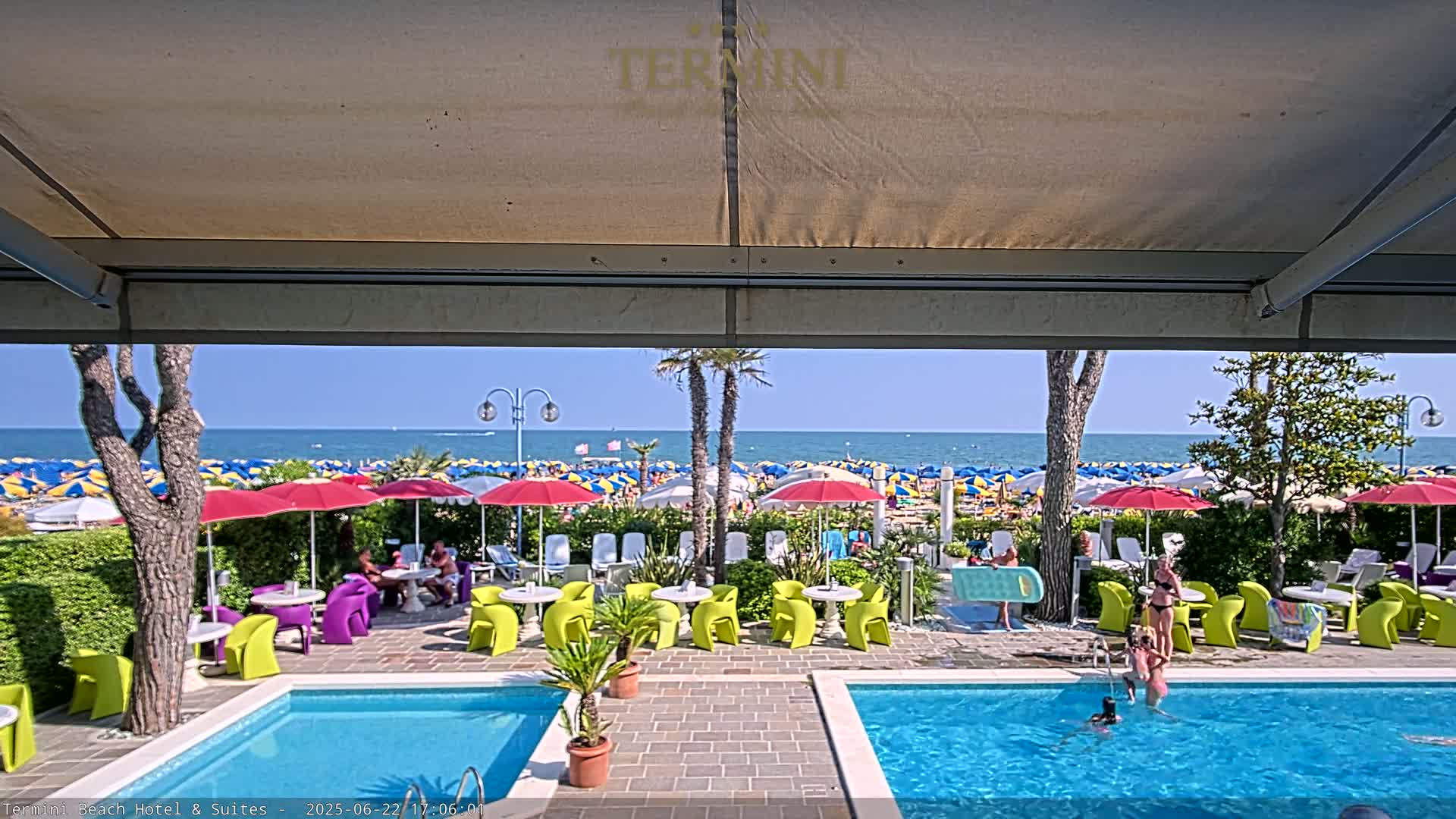 A sunny outdoor scene shows a resort pool area with bright green and purple chairs and tables, and a view of a crowded beach with many colorful umbrellas under a clear blue sky.