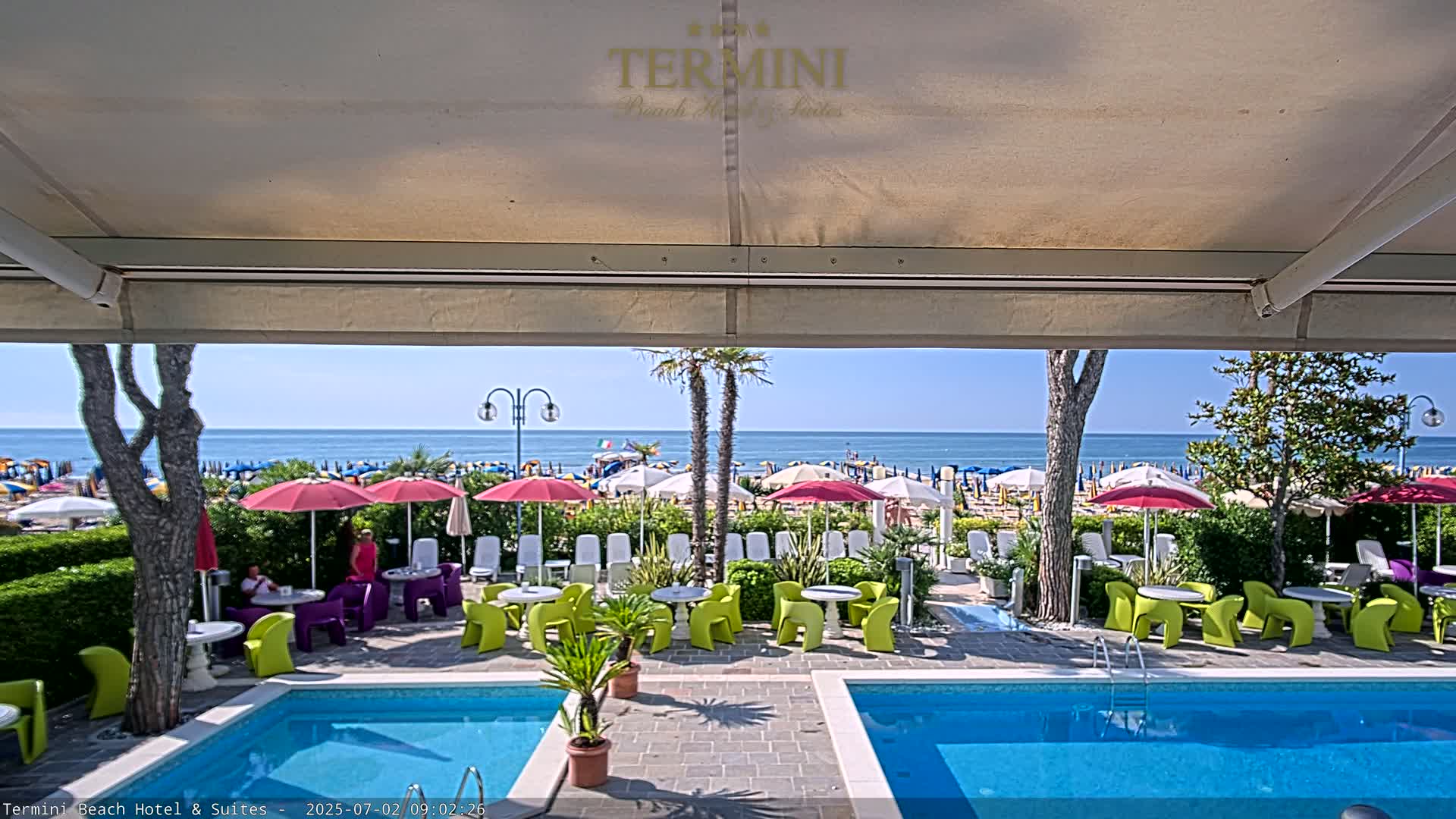 A sunny outdoor scene shows two swimming pools and patio furniture under a covered area, overlooking a beach with numerous umbrellas and a calm ocean.