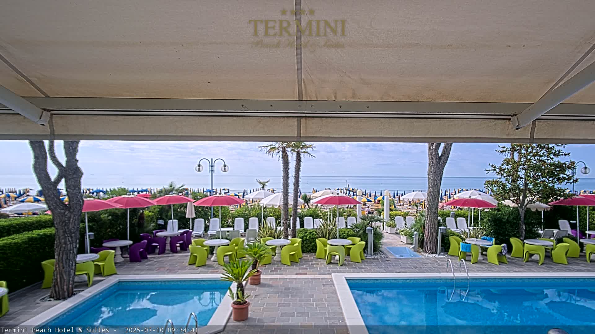 A view from under a patio awning shows a swimming pool, patio furniture, and a beach with numerous umbrellas under a mostly clear sky.