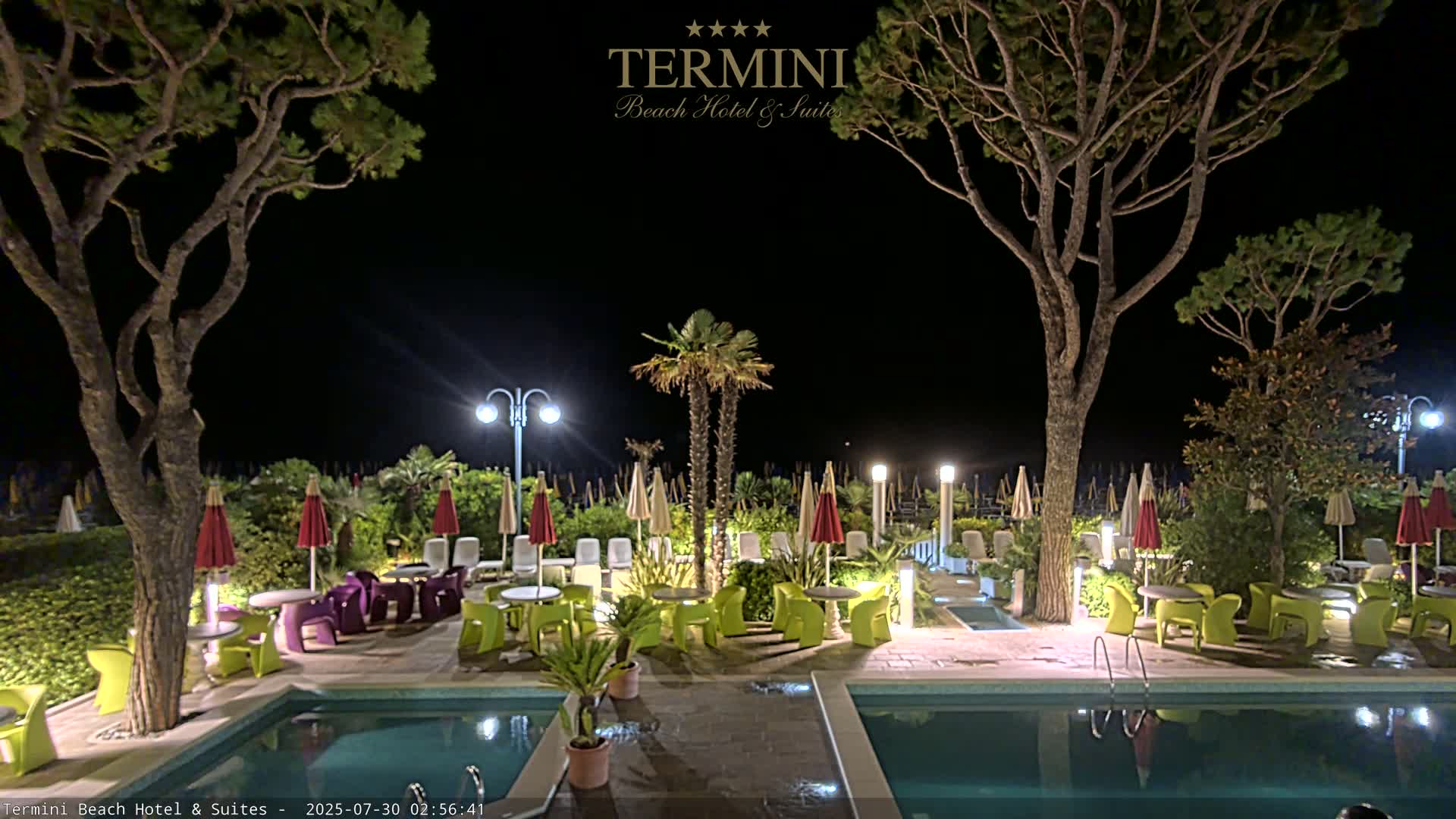 A view from under a patio awning shows a swimming pool, patio furniture, and a beach with numerous umbrellas under a mostly clear sky.