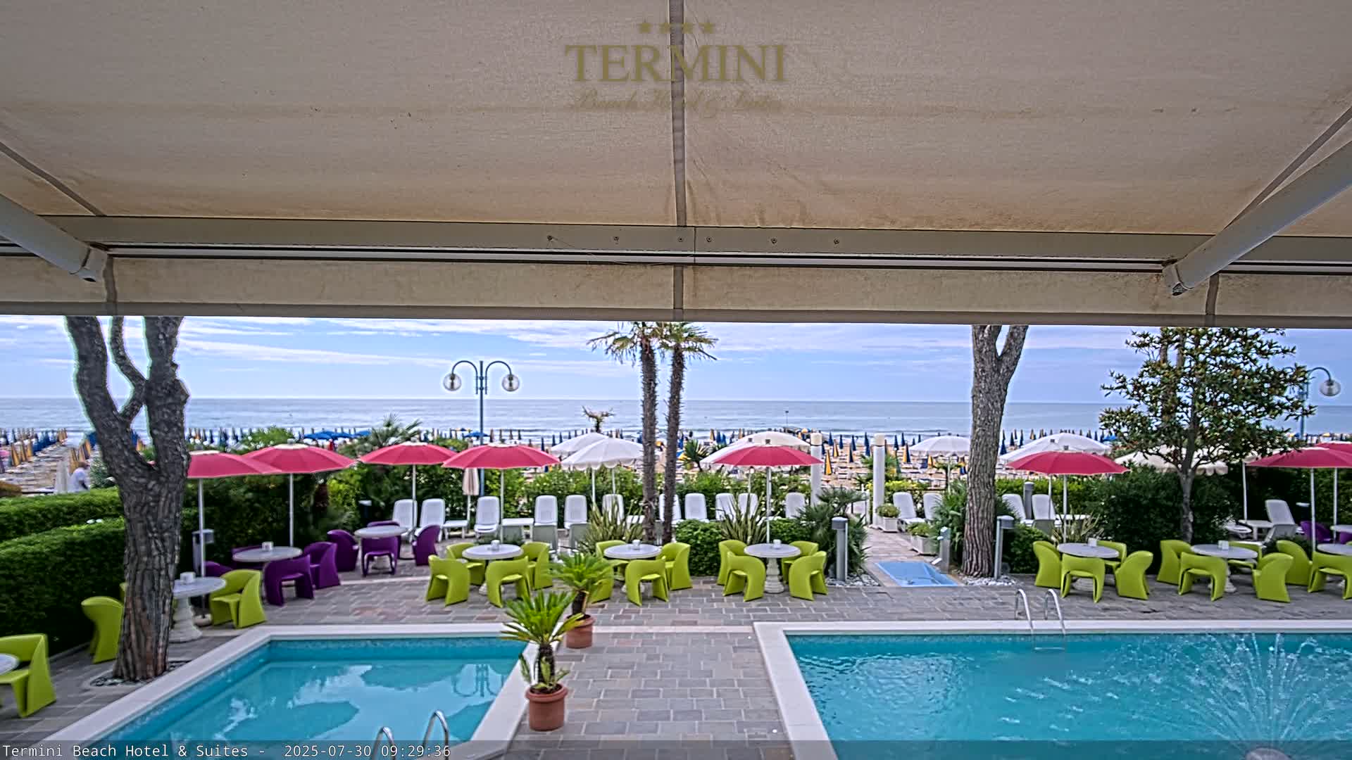 A view from under a patio awning shows a swimming pool, patio furniture, and a beach with numerous umbrellas under a partly cloudy sky.