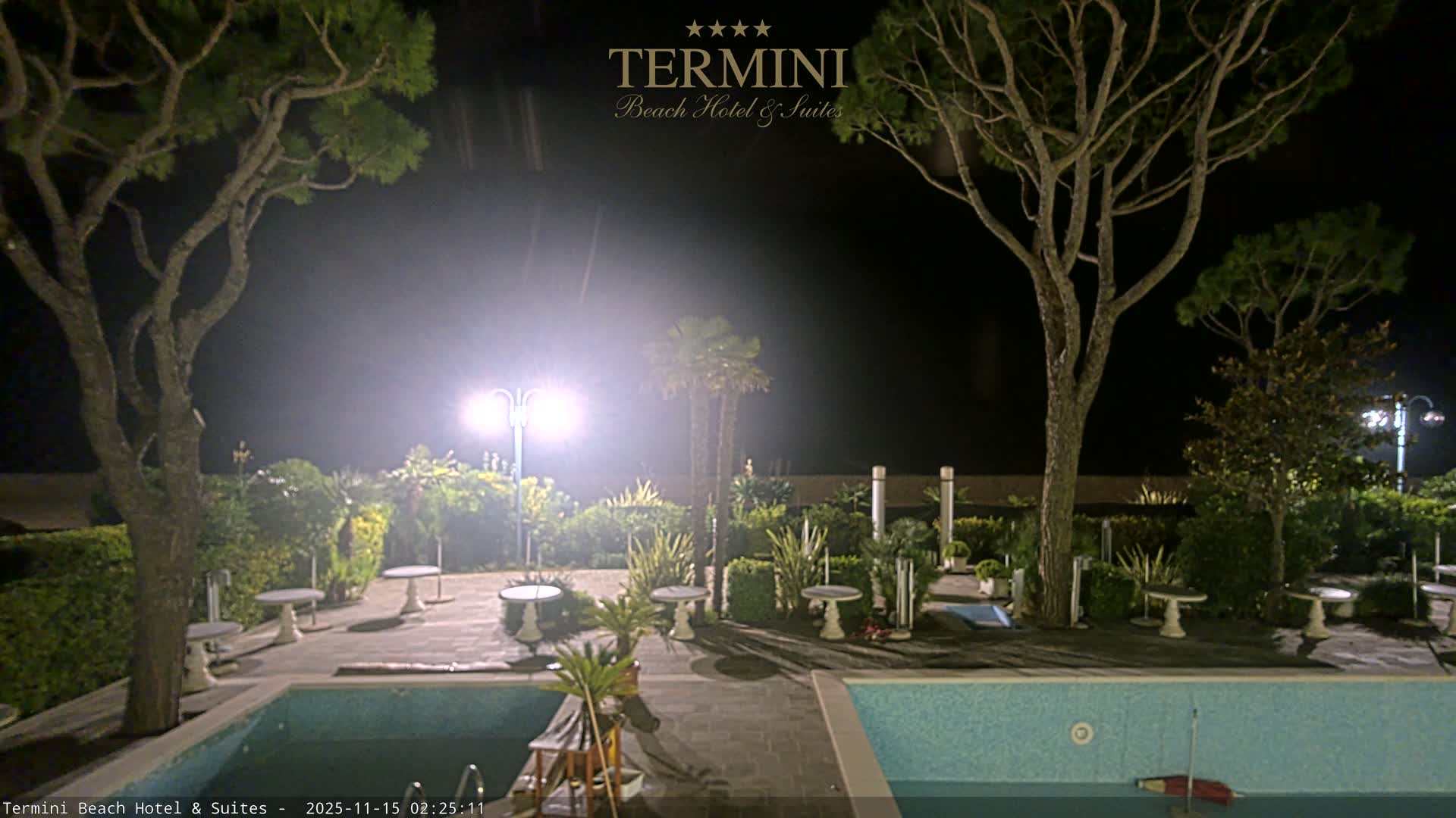 The image shows an outdoor resort area illuminated at night under clear skies, featuring two swimming pools, various trees including pines and palms, lush green landscaping, and several small tables on paved patios.