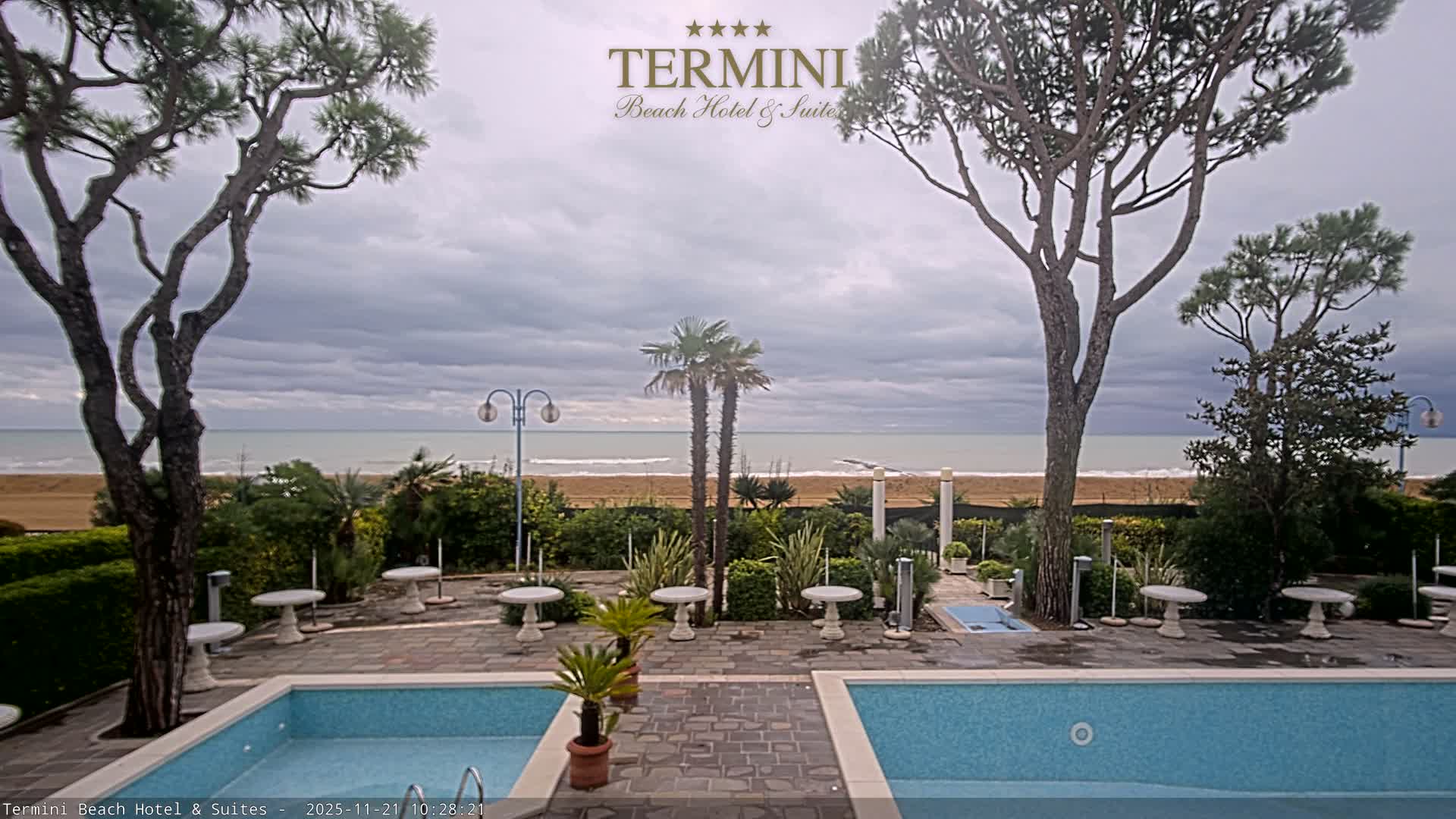 An overcast day reveals a coastal scene featuring two swimming pools and a patio with white tables in the foreground, backed by lush greenery, a sandy beach, and the ocean under a grey, cloudy sky.