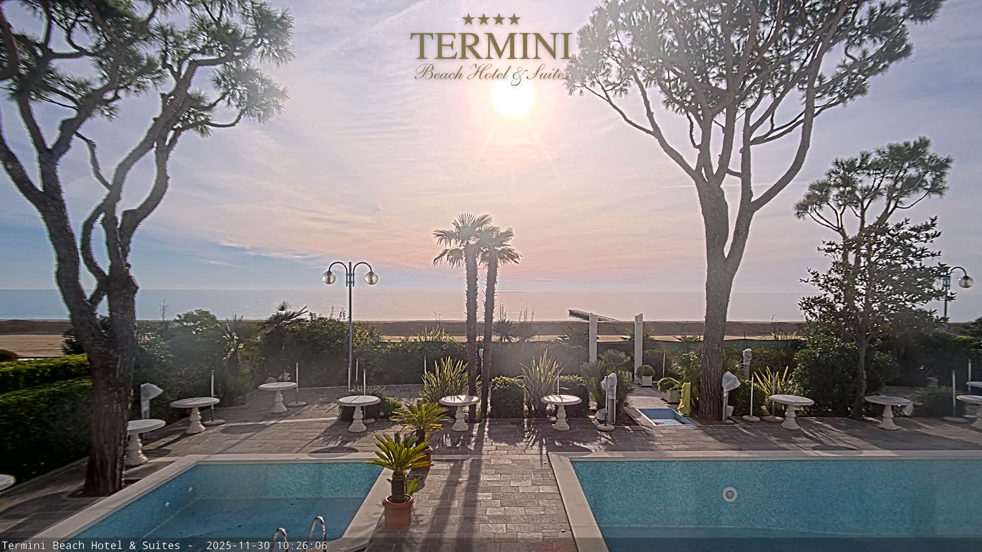 Under a clear, sunny sky with warm hues, a tranquil beach resort scene features two swimming pools, patio tables, and lush trees overlooking the calm ocean and sandy beach.