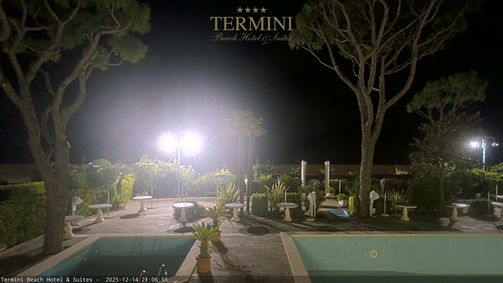 Under a clear, sunny sky with warm hues, a tranquil beach resort scene features two swimming pools, patio tables, and lush trees overlooking the calm ocean and sandy beach.