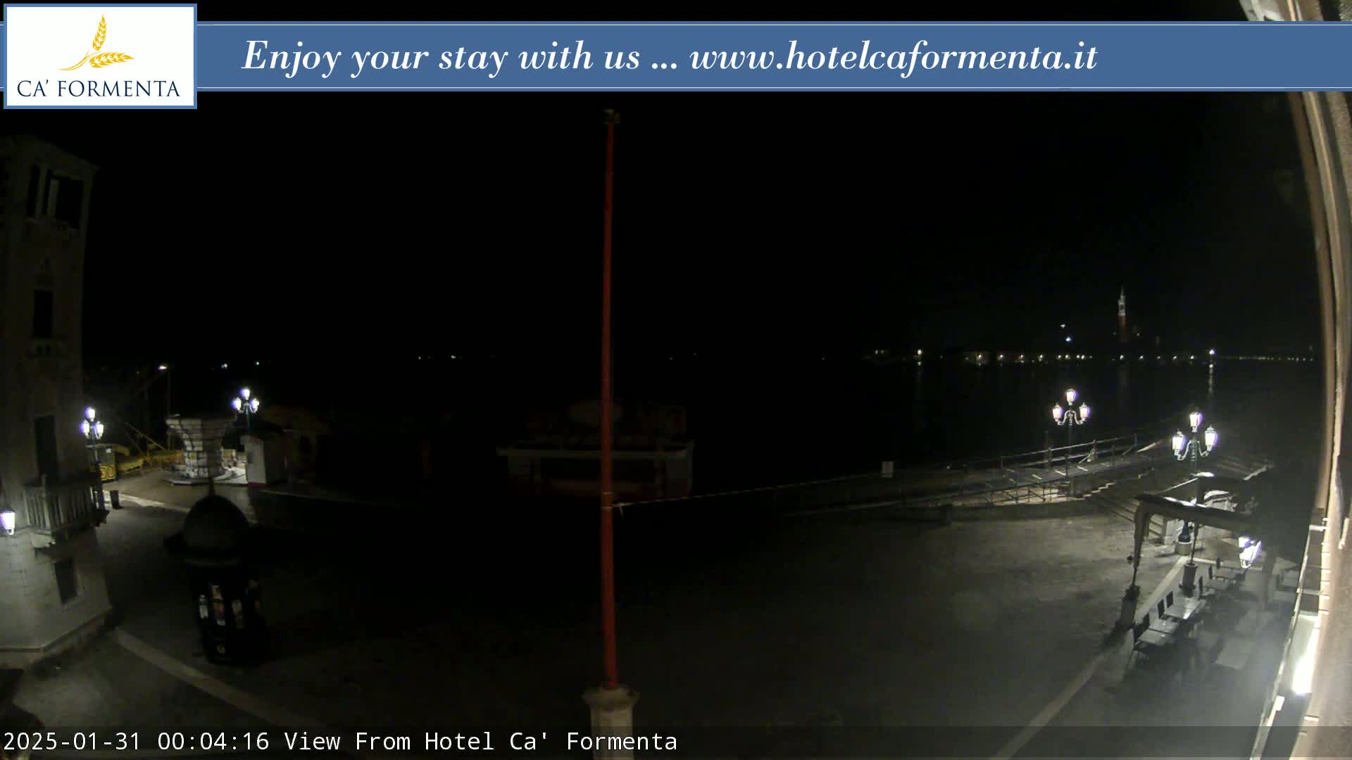 A nighttime view of a waterfront plaza shows a calm body of water with lights visible across it.
