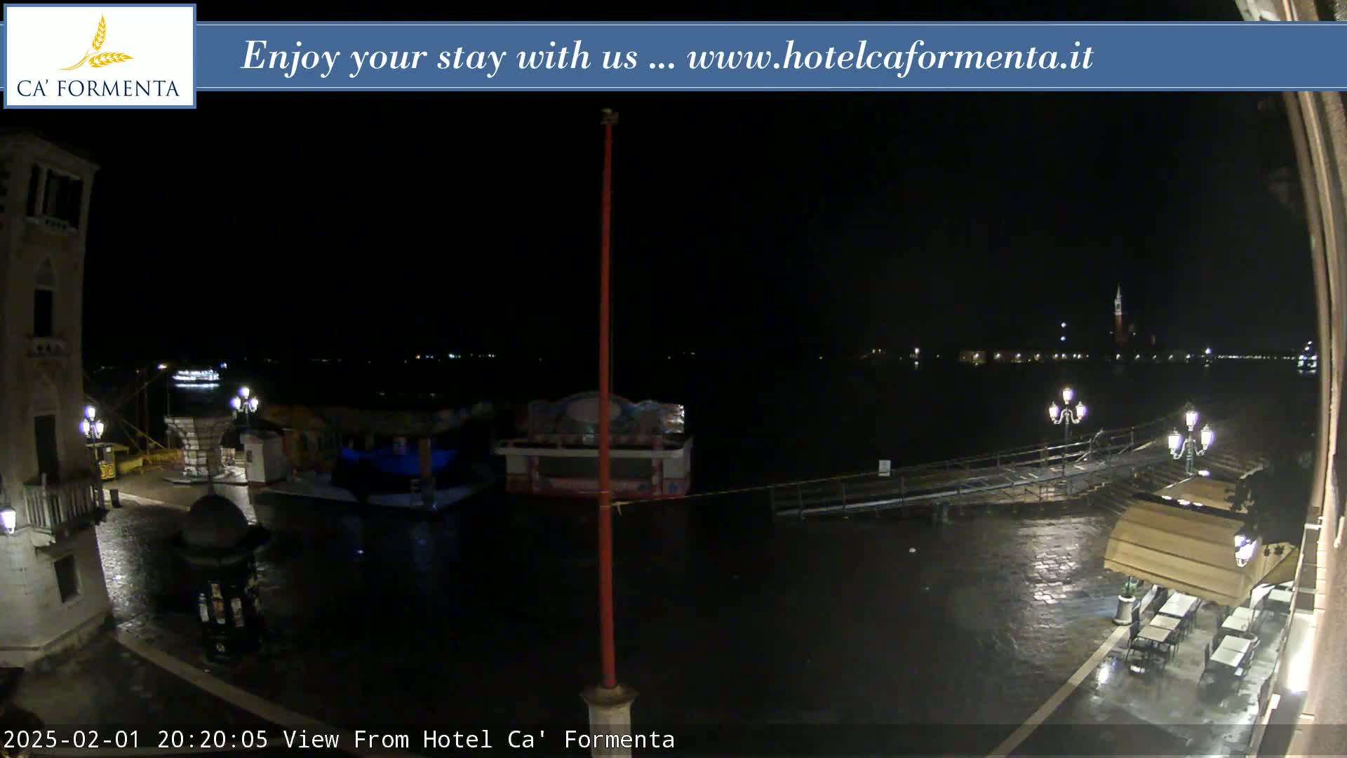 A nighttime view of a waterfront area shows a wet cobblestone plaza with a few outdoor tables and chairs, a canal with boats and lights across the water, and a tall flagpole in the foreground.