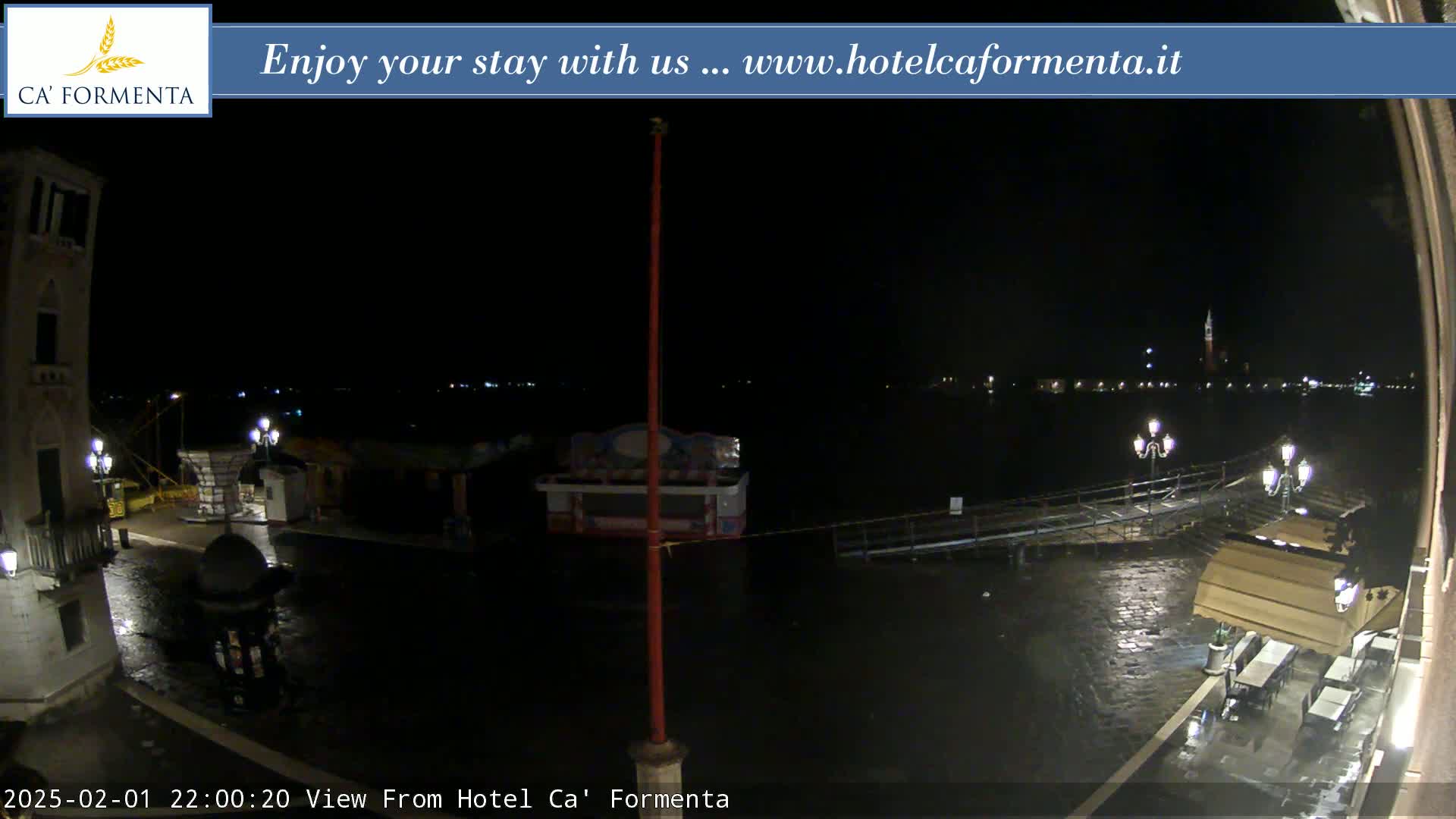 A nighttime view of a waterfront plaza, showing dark water and a few illuminated buildings and outdoor dining areas in the rain.