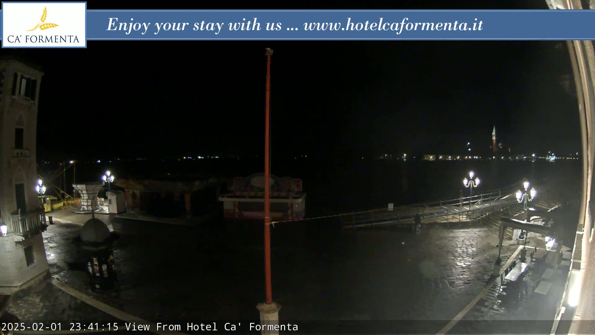 A nighttime view of a waterfront plaza, wet from rain, shows a few people, several lights, and buildings across the water.