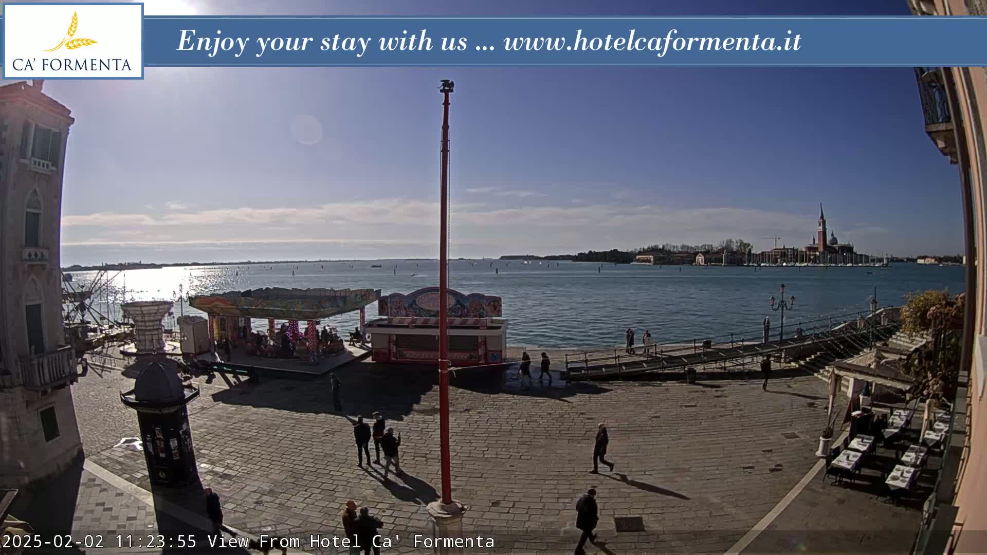A sunny day overlooks a paved plaza with a small carousel and a few people walking, beside a calm body of water and a distant island.
