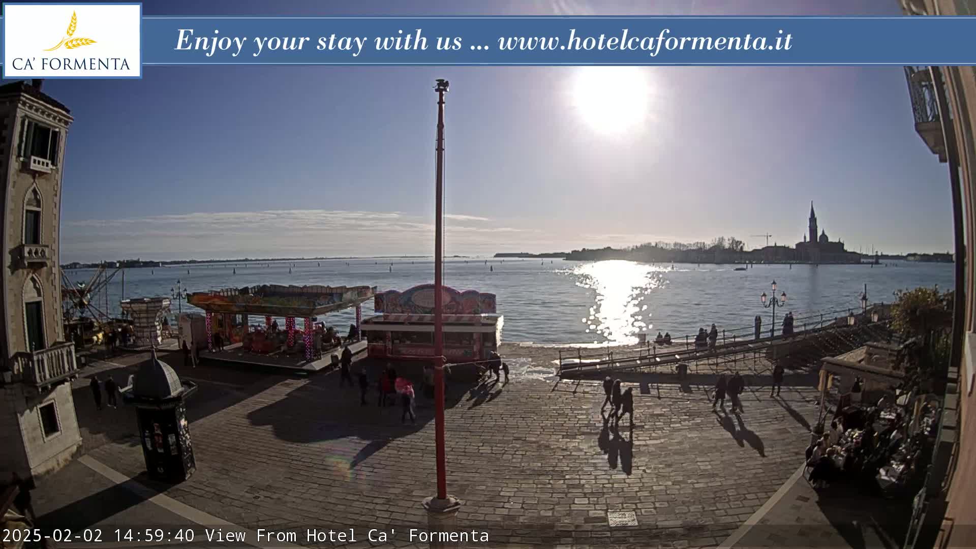 A sunny waterfront plaza scene shows a fairground ride, people walking, and a distant view of a city across the water.