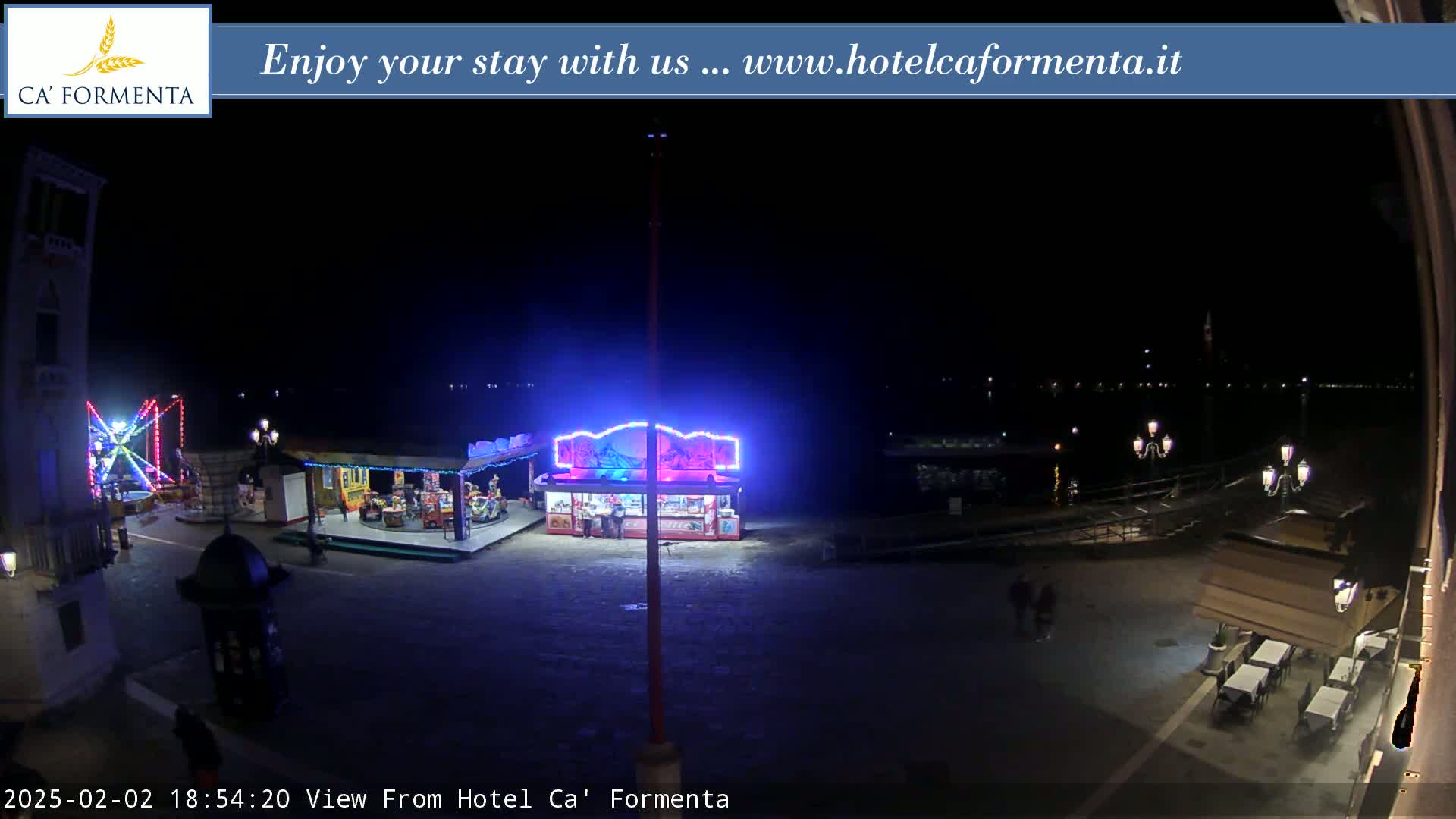 A nighttime view of a town square with illuminated carnival rides, food stands, and outdoor seating at restaurants near a body of water.
