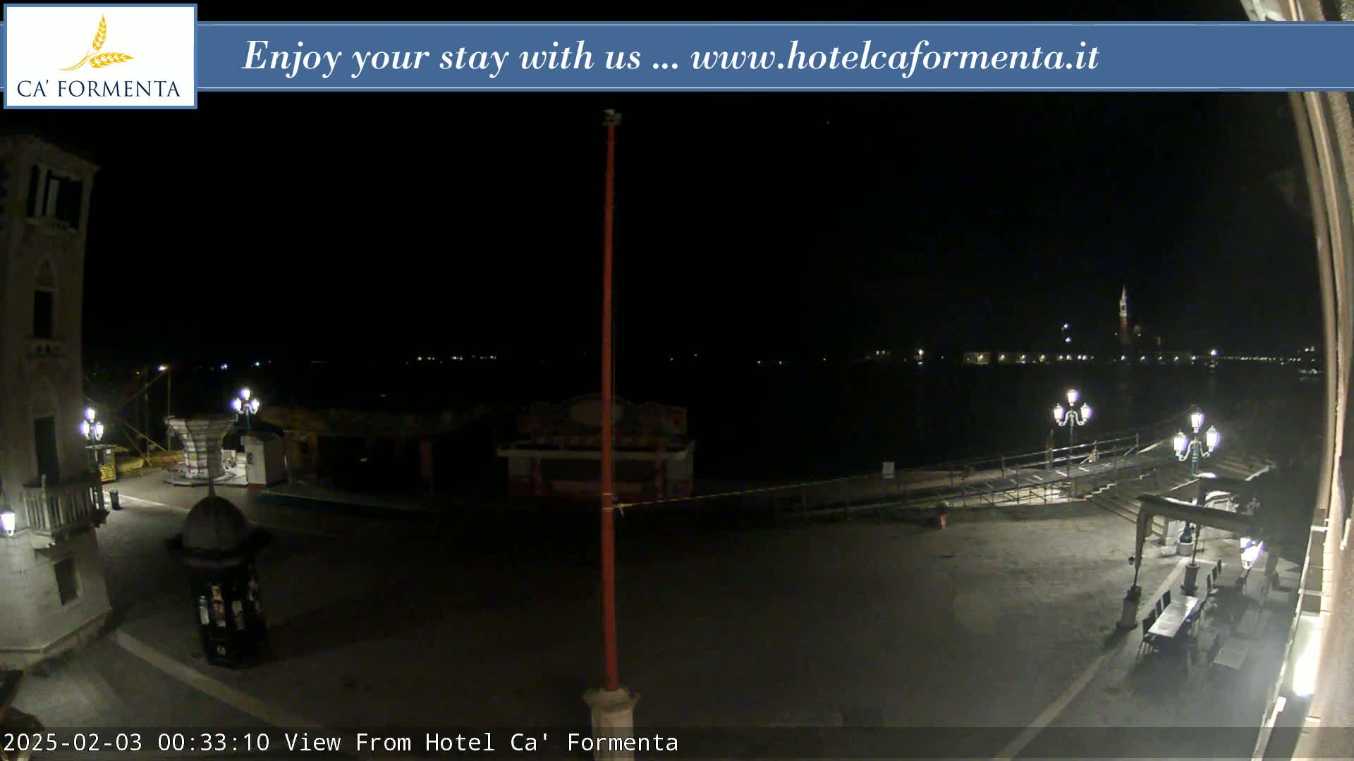 A nighttime view of a plaza overlooking a dark body of water with faintly visible lights across it, under a clear, dark sky.