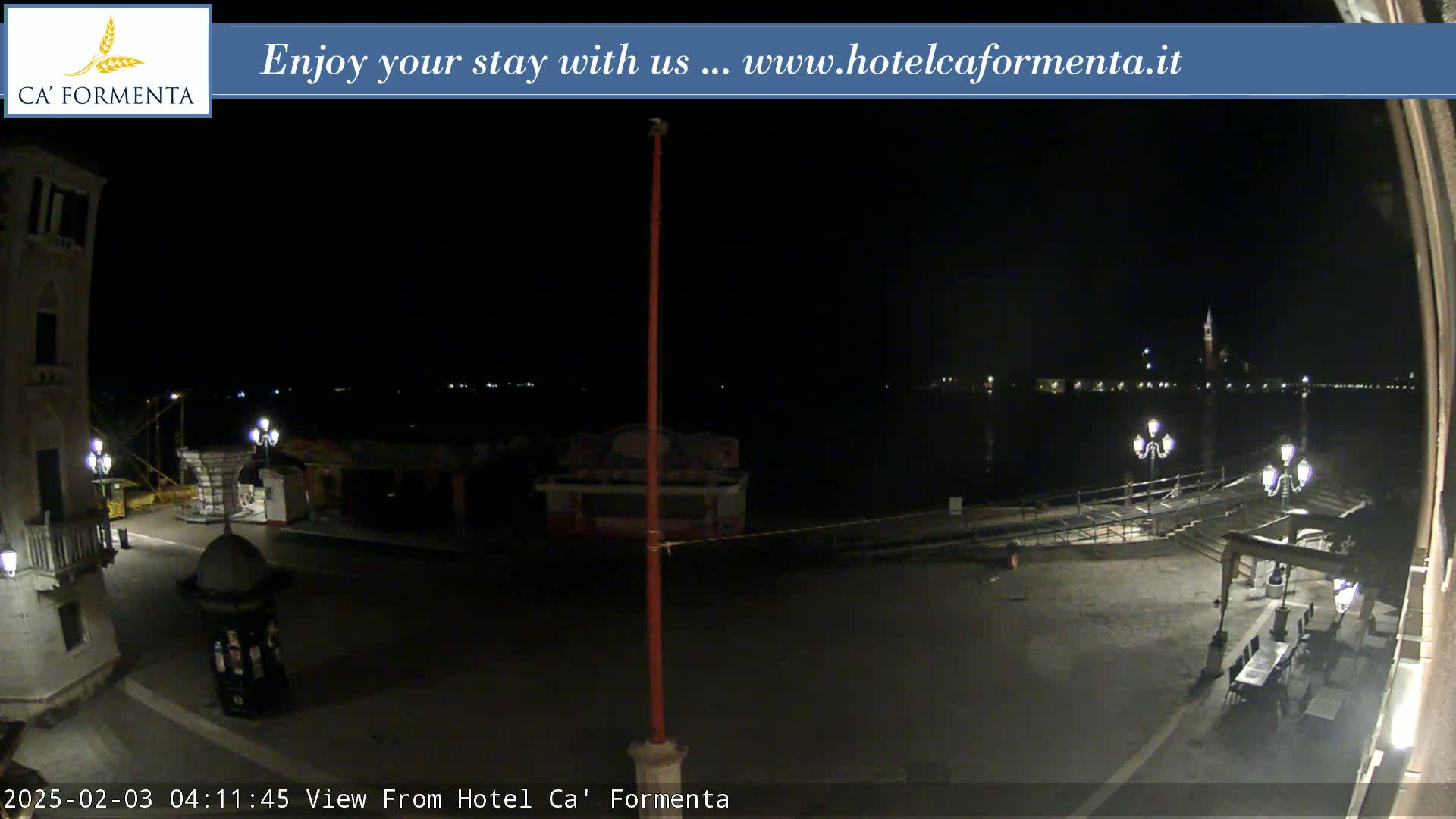 A nighttime view of a waterfront plaza with a calm body of water and distant lights across it.