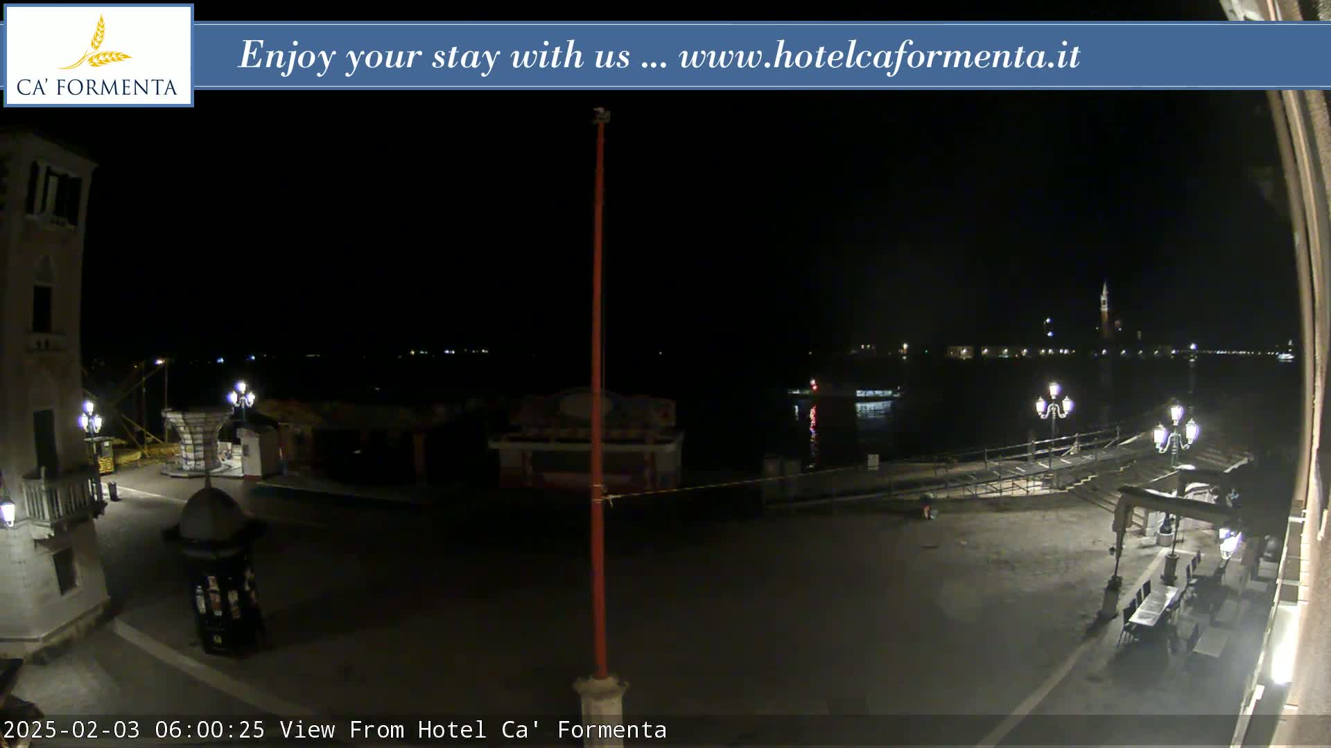 A nighttime view of a waterfront plaza with calm water, distant lights, and a few streetlamps.
