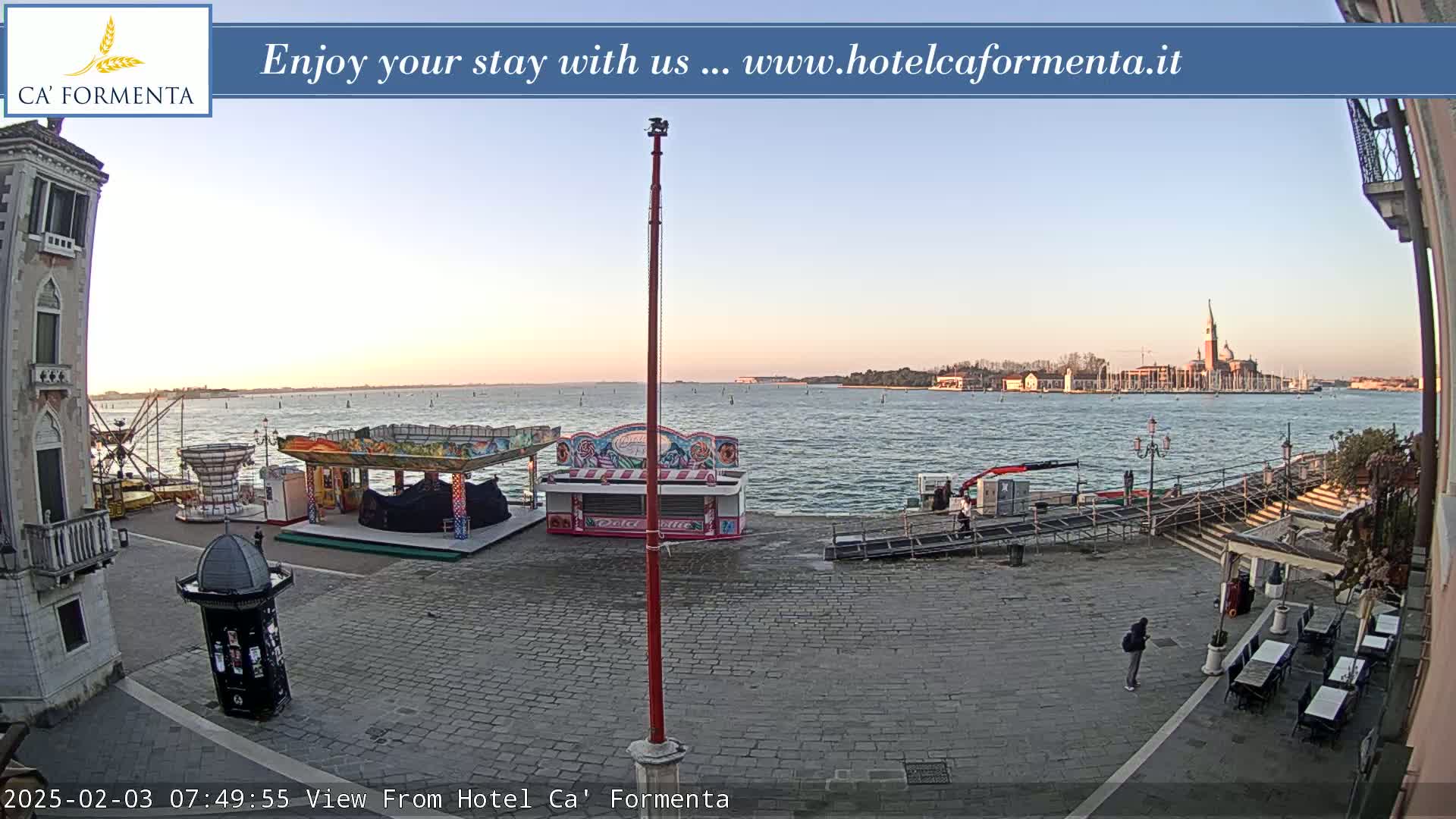 A calm, dawn-lit waterfront square features several carnival rides and an outdoor cafe, with a distant view of a city across the water.