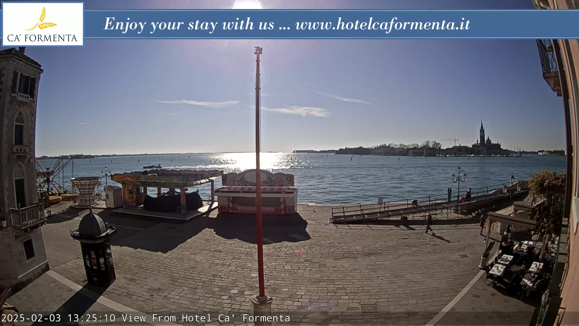 A sunny waterfront view shows a paved plaza with several small kiosks and a distant cityscape across the water.