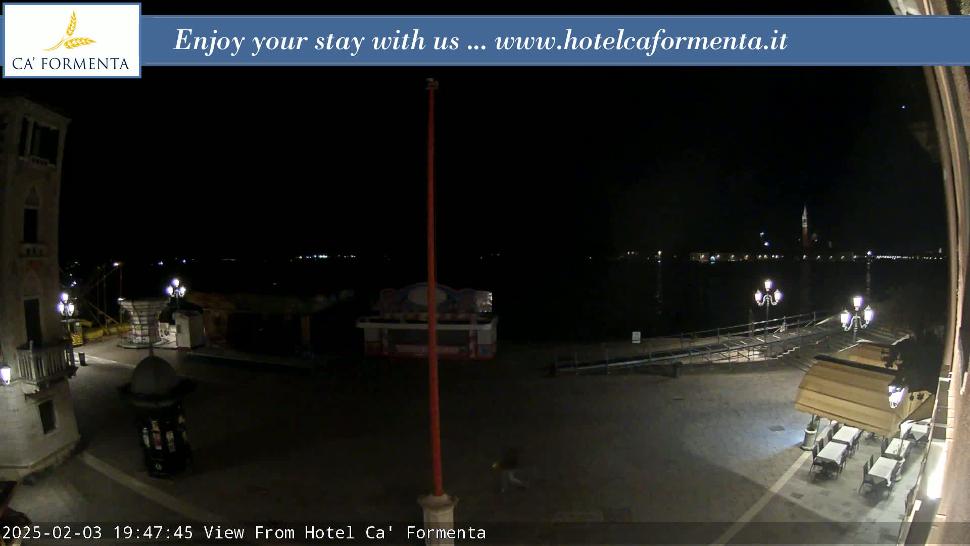 A nighttime view of a plaza near a body of water, with a few outdoor lights and a small structure in the foreground.