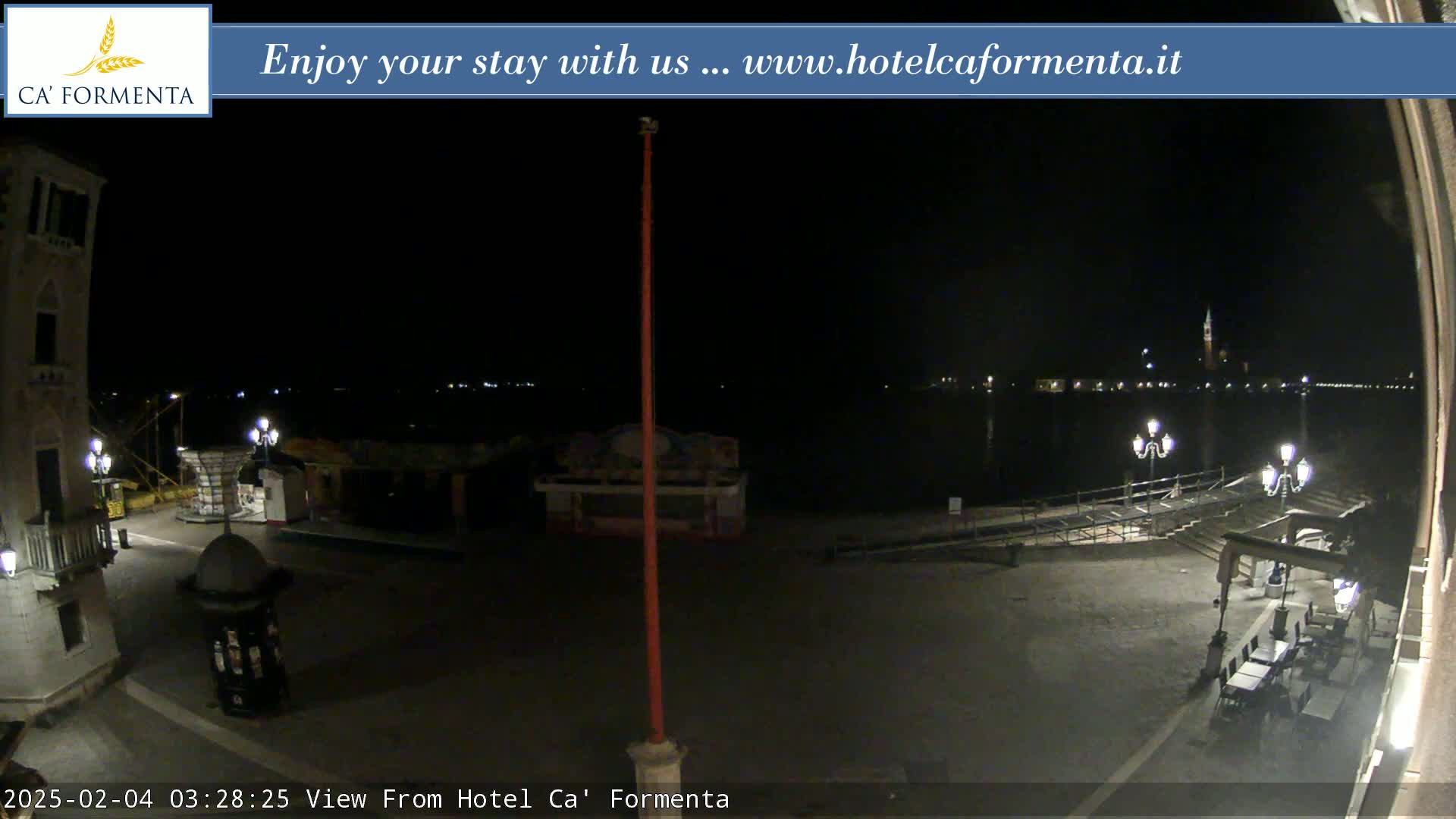 A nighttime view of a waterfront plaza with dimly lit buildings and a body of water showing distant lights across it.