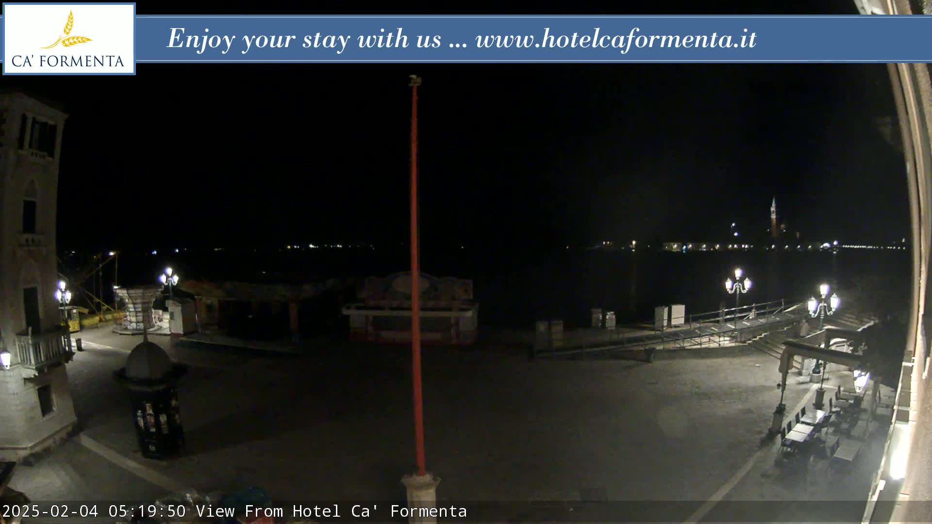 A nighttime view of a waterfront plaza with sparsely lit buildings and a dark body of water in the background.