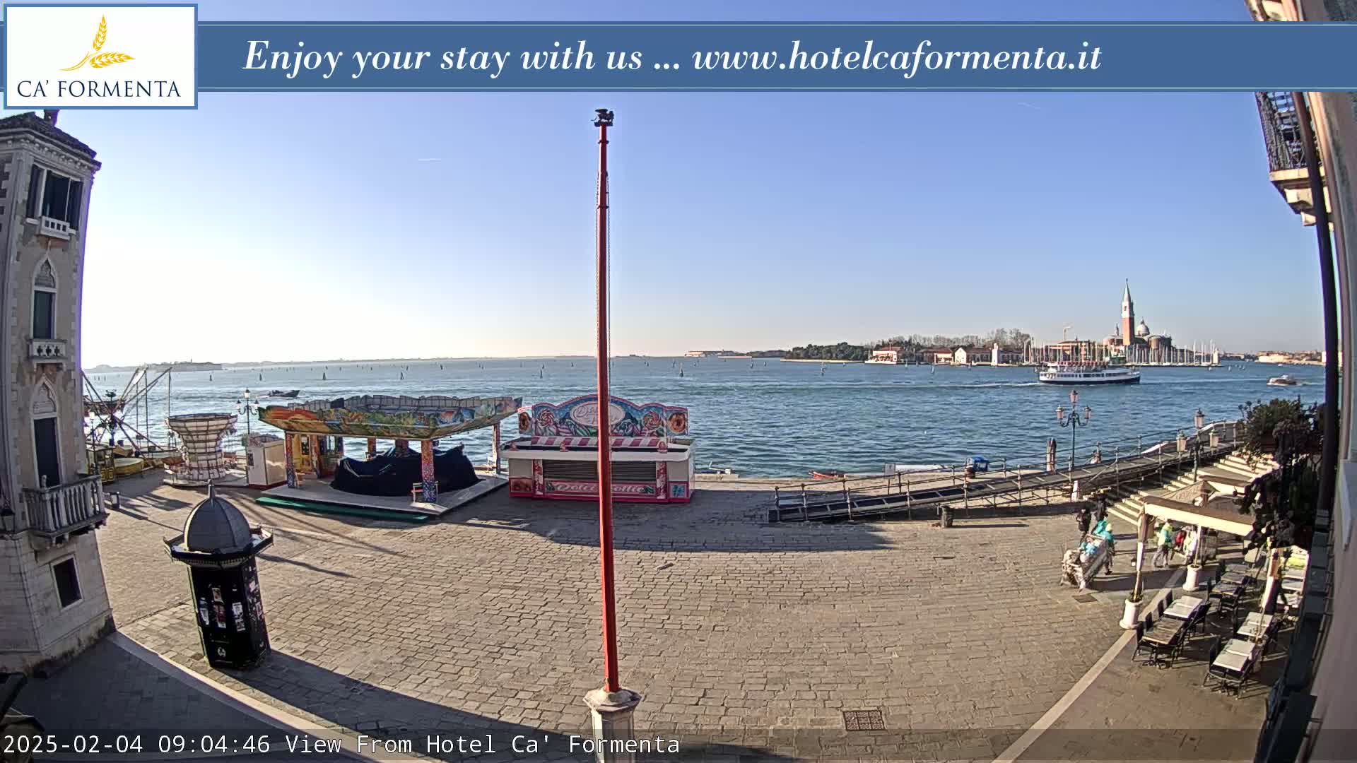 A sunny day reveals a cobblestone plaza with several closed carnival game booths, a waterway with a passing boat and distant buildings, and a few people near outdoor seating.