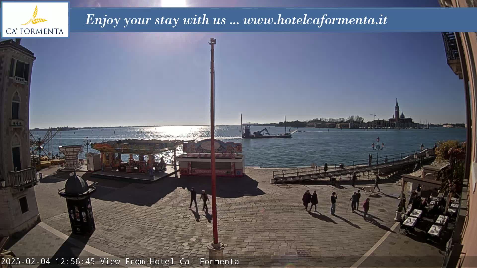 A sunny day overlooking a calm body of water with a small amusement park and several people walking on a paved area near a building.