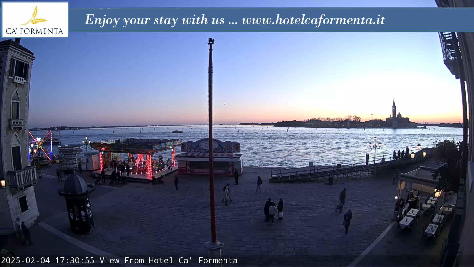 A twilight view shows a waterfront plaza with a small amusement park, several people, and a distant cityscape across calm water.