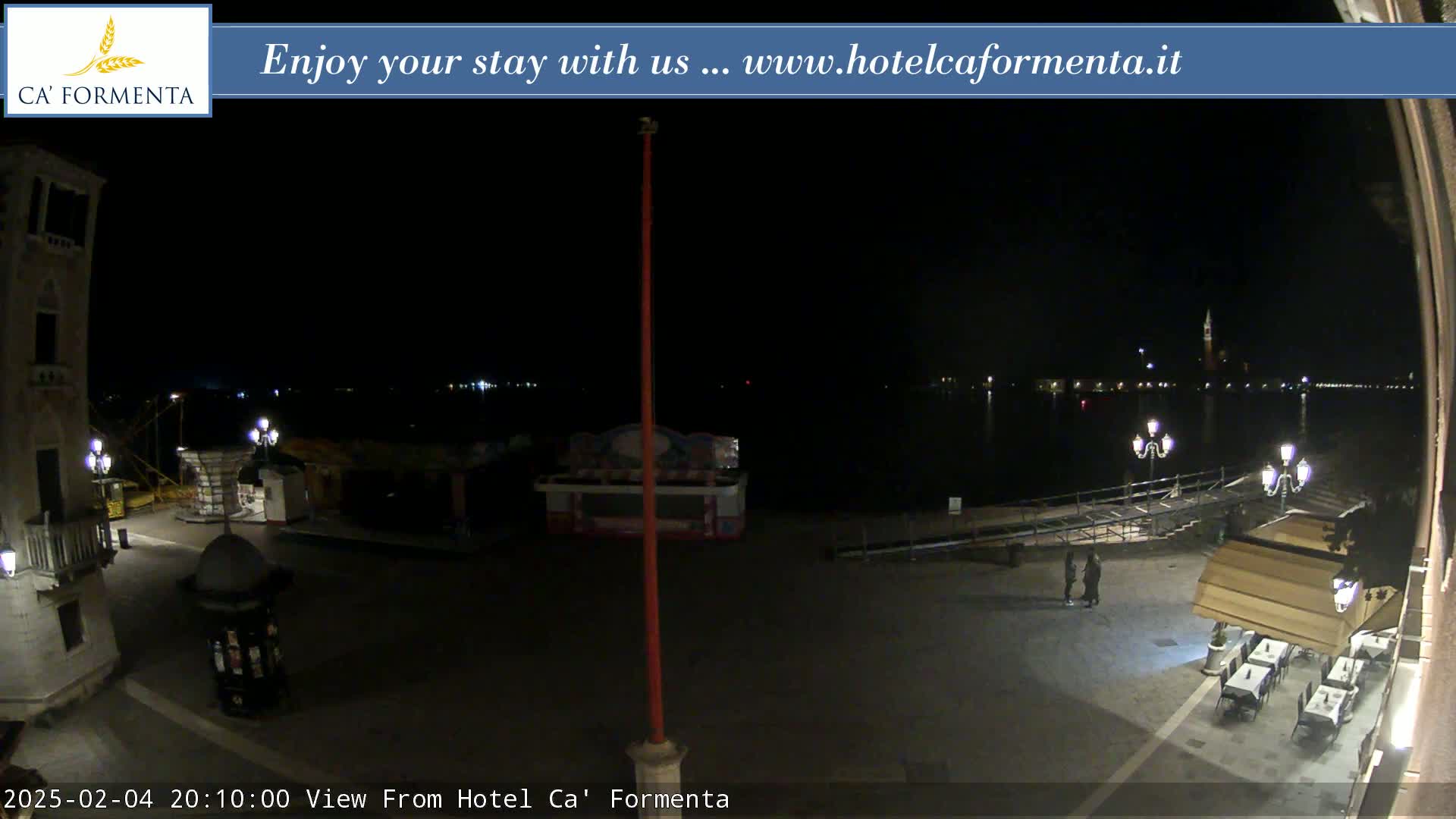 A nighttime view of a waterfront plaza with a calm body of water and distant lights across it, some outdoor seating at a restaurant, and a few people walking by.