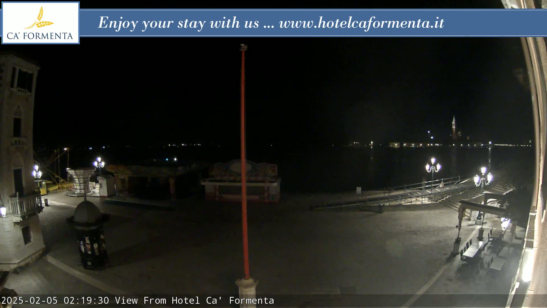 A nighttime view of a waterfront plaza with several lamps, some outdoor furniture, and a body of water with distant lights across it.