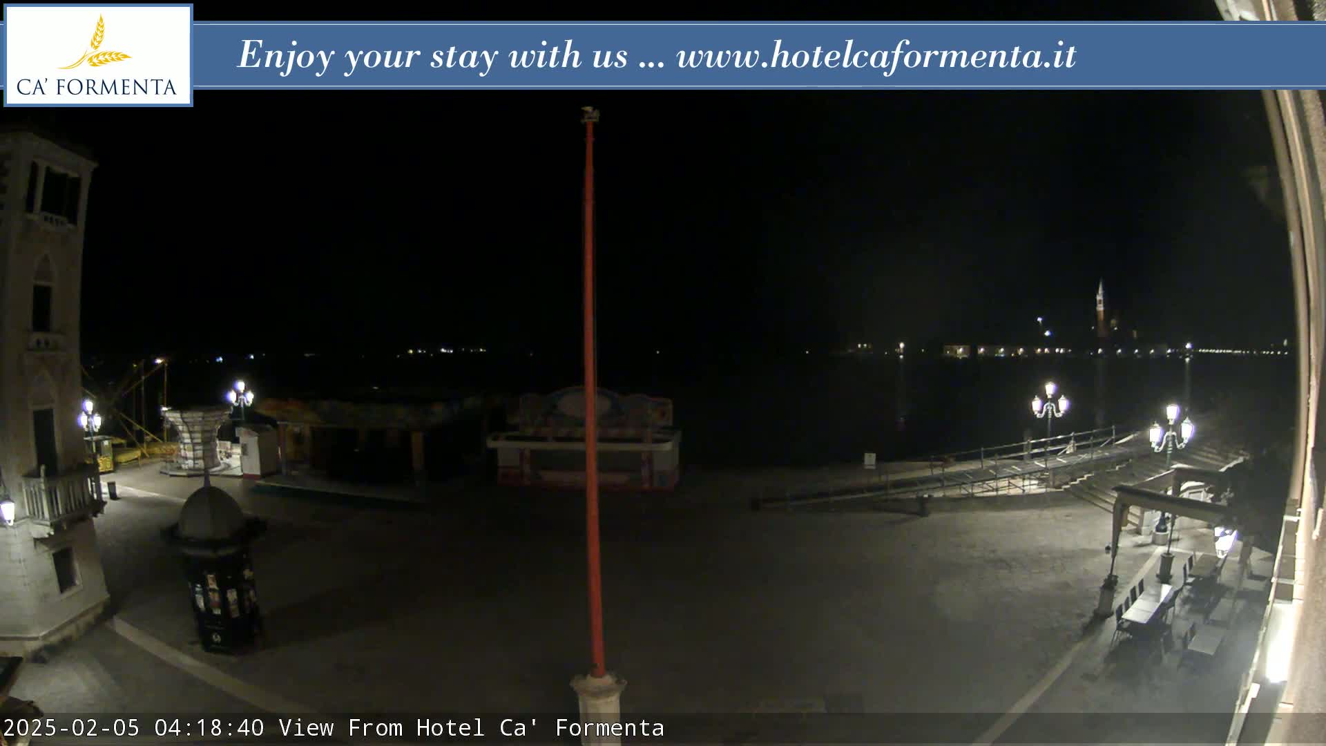 A nighttime view of a plaza and waterfront, with a few lights illuminating the scene and calm water in the background.