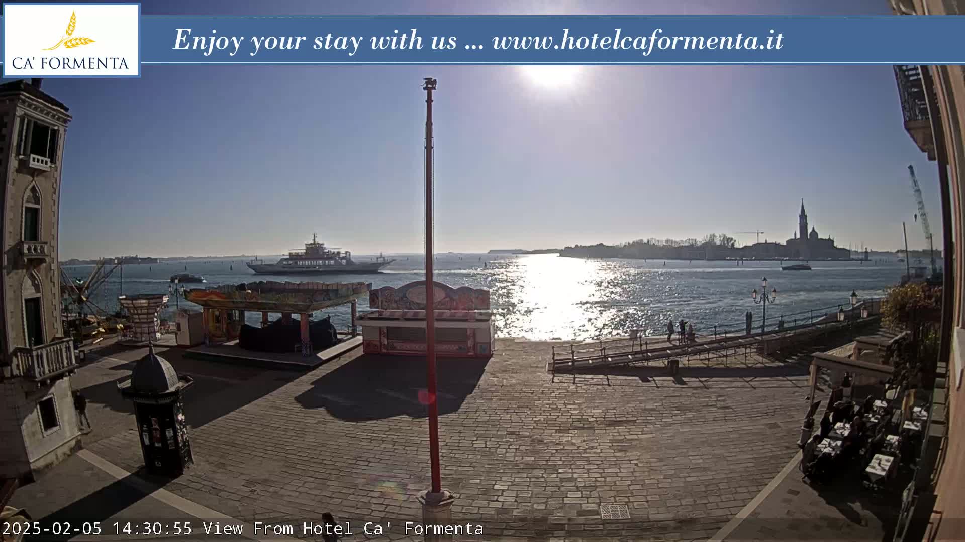 A sunny waterfront scene shows a paved plaza with small kiosks and a distant view of water, ships, and a city skyline.