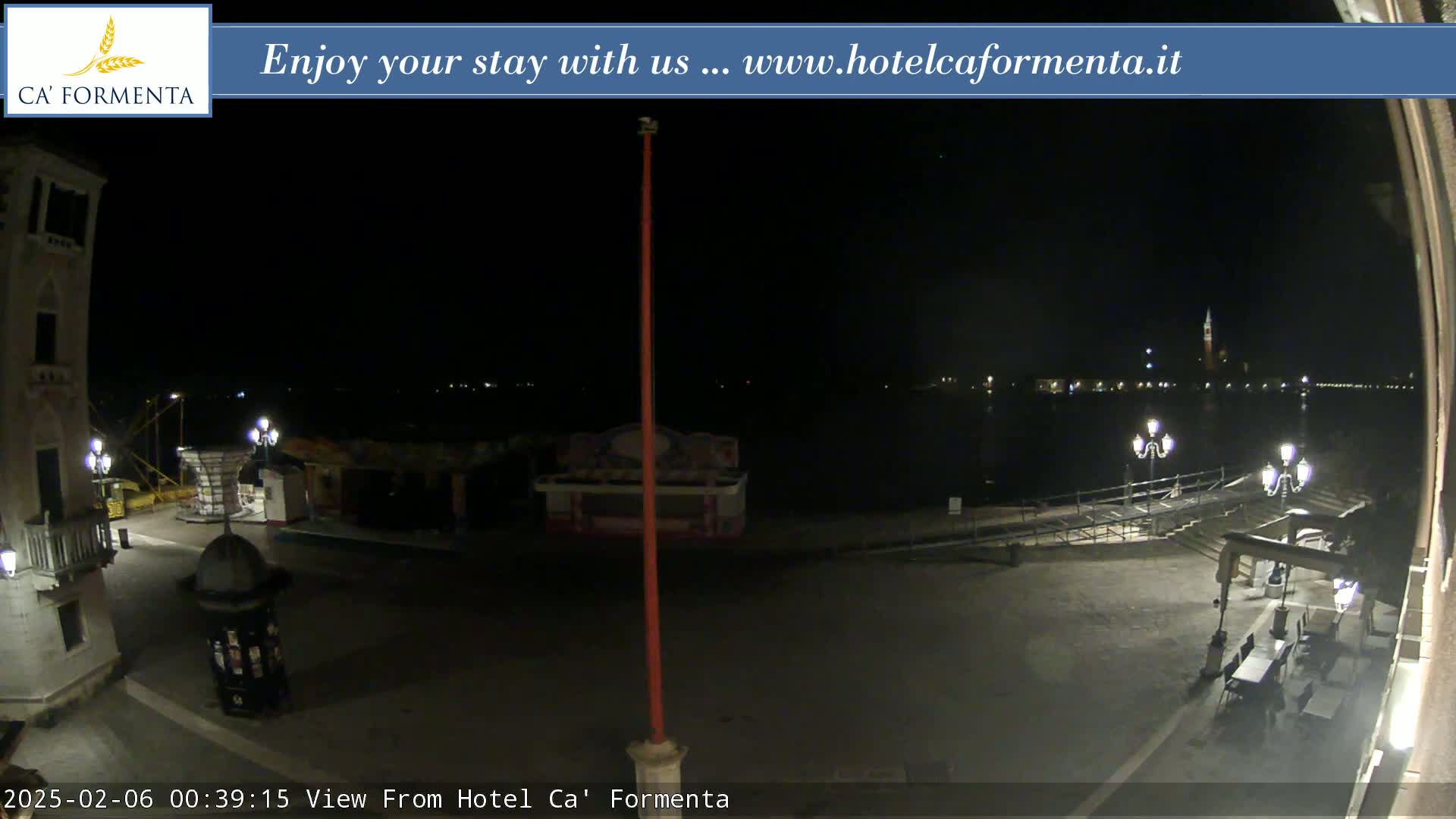 A nighttime view of a waterfront plaza with a body of water and distant city lights, under clear skies.