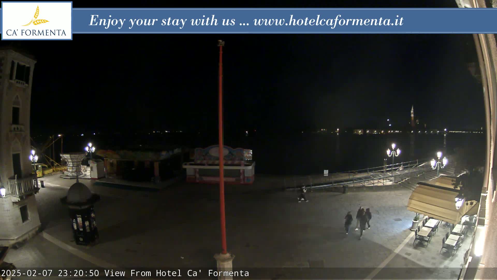 A nighttime view of a waterfront square with a few people walking, some carnival-style stalls, and a body of water with lights across it in the distance.
