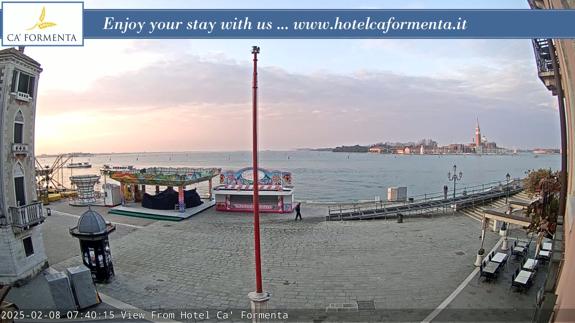 A waterfront plaza with a few carnival-style food stalls and a calm body of water showing a distant cityscape under a light, cloudy sky at dawn.