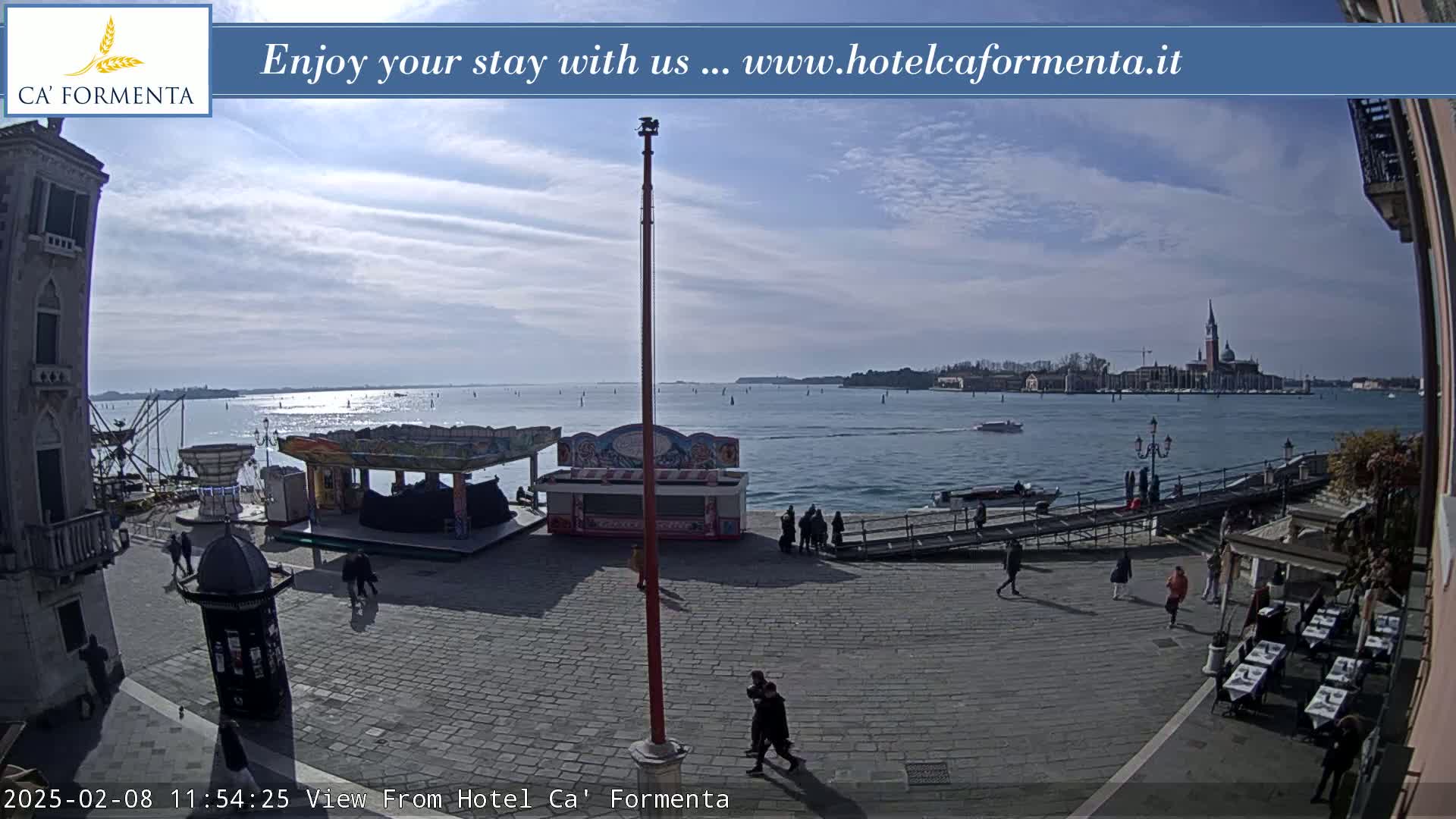 A sunny day shows a waterfront paved plaza with a few people walking, some small food stalls and a distant view of a city across calm water.