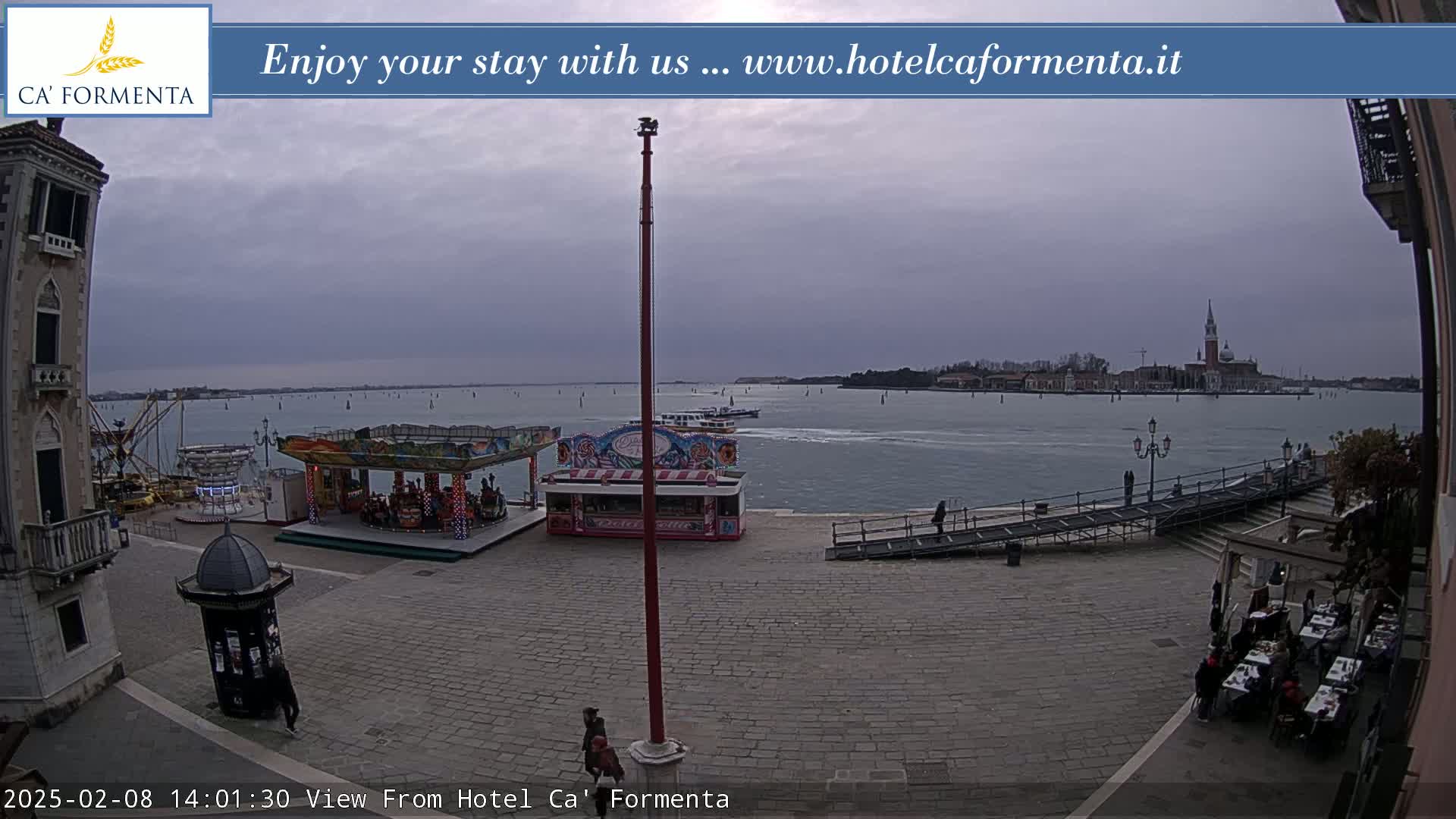 A view of a waterfront plaza with a carousel and other fairground attractions, overlooking calm water and distant buildings under an overcast sky.