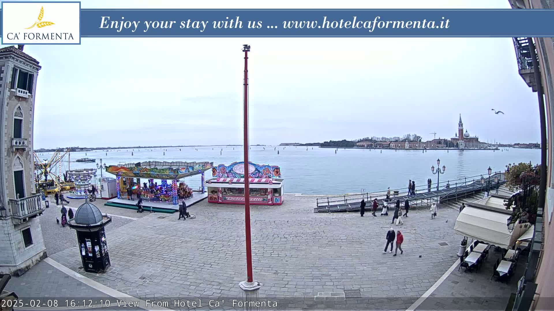 A paved plaza with a carousel and food stand overlooks a calm body of water, with a distant cityscape and overcast sky.
