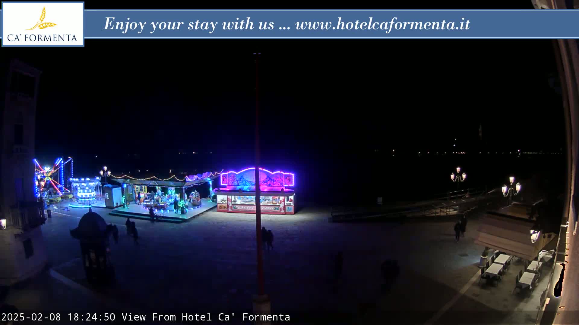 A nighttime view of a brightly lit carnival area near a body of water, with a few people walking around.