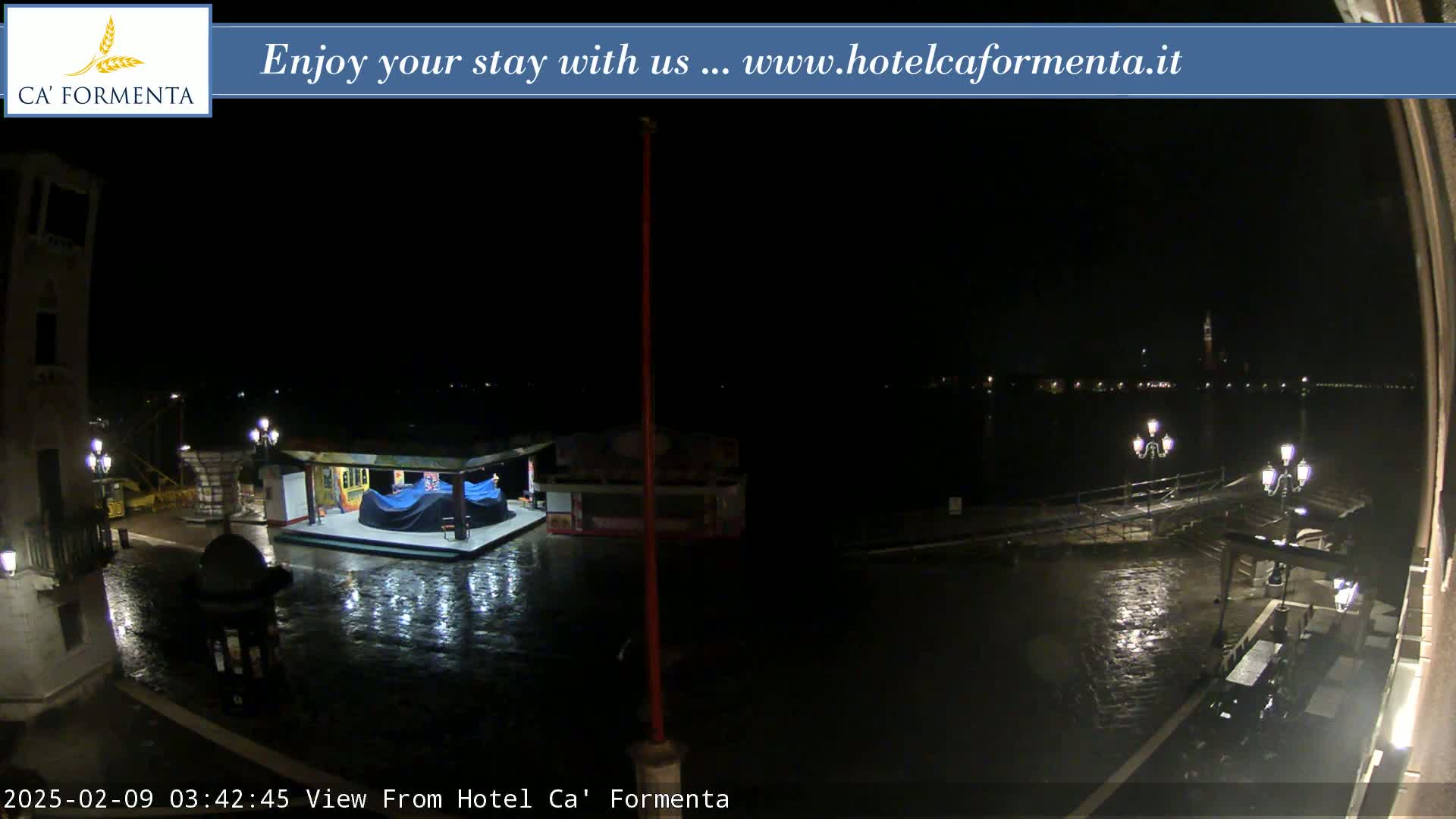 A nighttime, rain-slicked waterfront scene shows a partially submerged stage and surrounding structures, with distant city lights across the water.