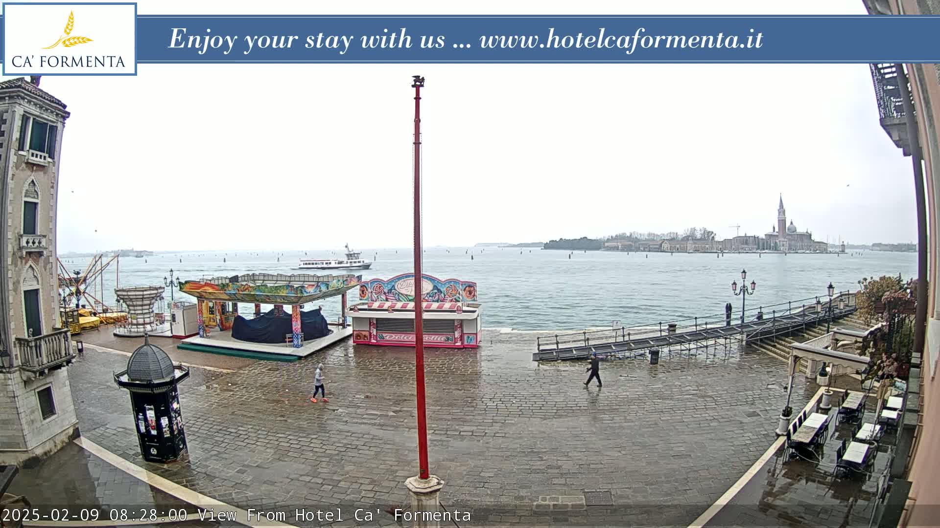 A wet cobblestone plaza overlooks a calm body of water with a distant island and several carnival-style kiosks.