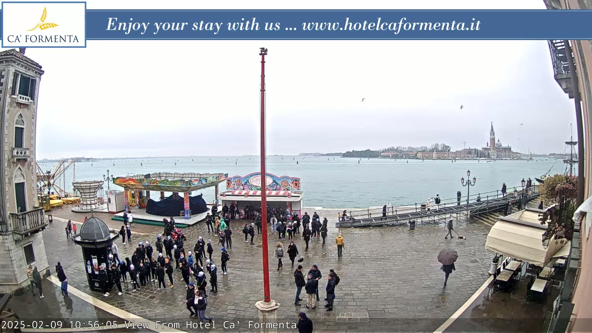 A wet, overcast day reveals a waterfront plaza with people gathered near carnival-style rides and buildings, overlooking a calm body of water and distant cityscape.