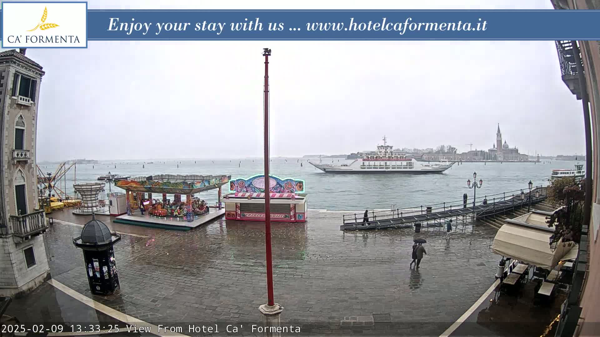 A ferry is visible on a waterway under an overcast sky, with a wet cobblestone plaza containing a small amusement park and several people walking nearby.
